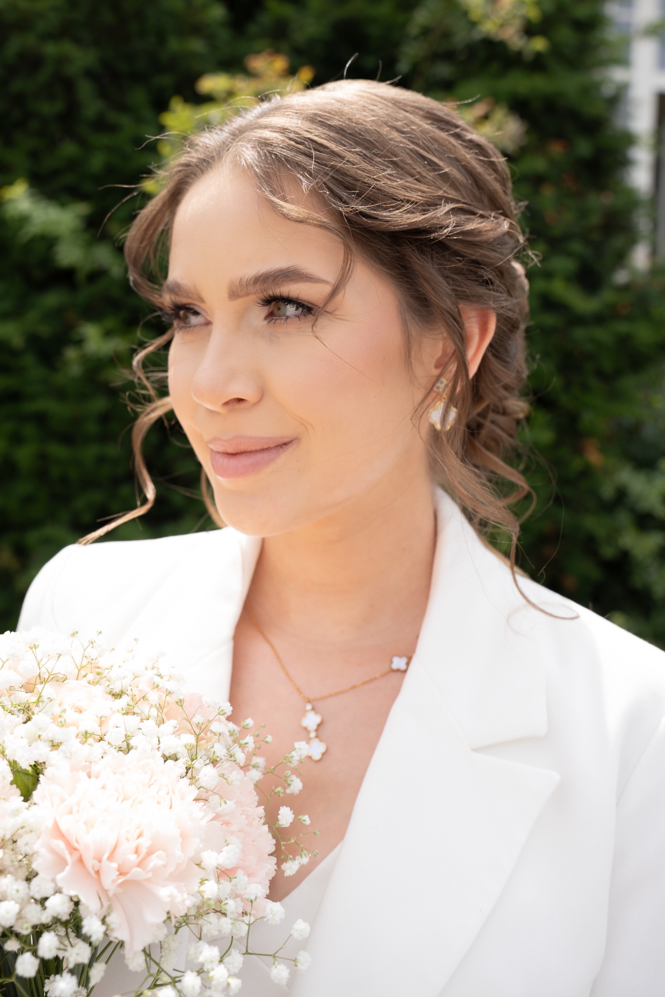 Portret kobiety w białym żakiecie, z delikatnym makijażem i upiętymi włosami, trzymającej bukiet bladoróżowych goździków i gipsówki na tle zieleni.