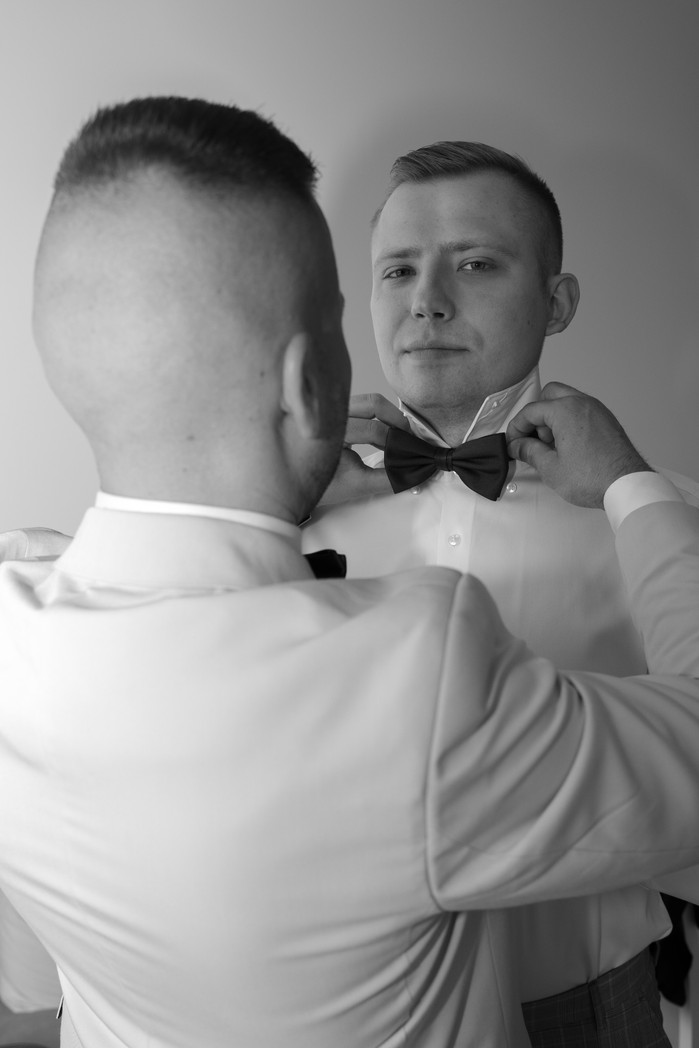 Czarno-białe zdjęcie mężczyzny dopinającego muchę drugiemu mężczyźnie w białej koszuli i marynarce, przygotowania ślubne w zbliżeniu.