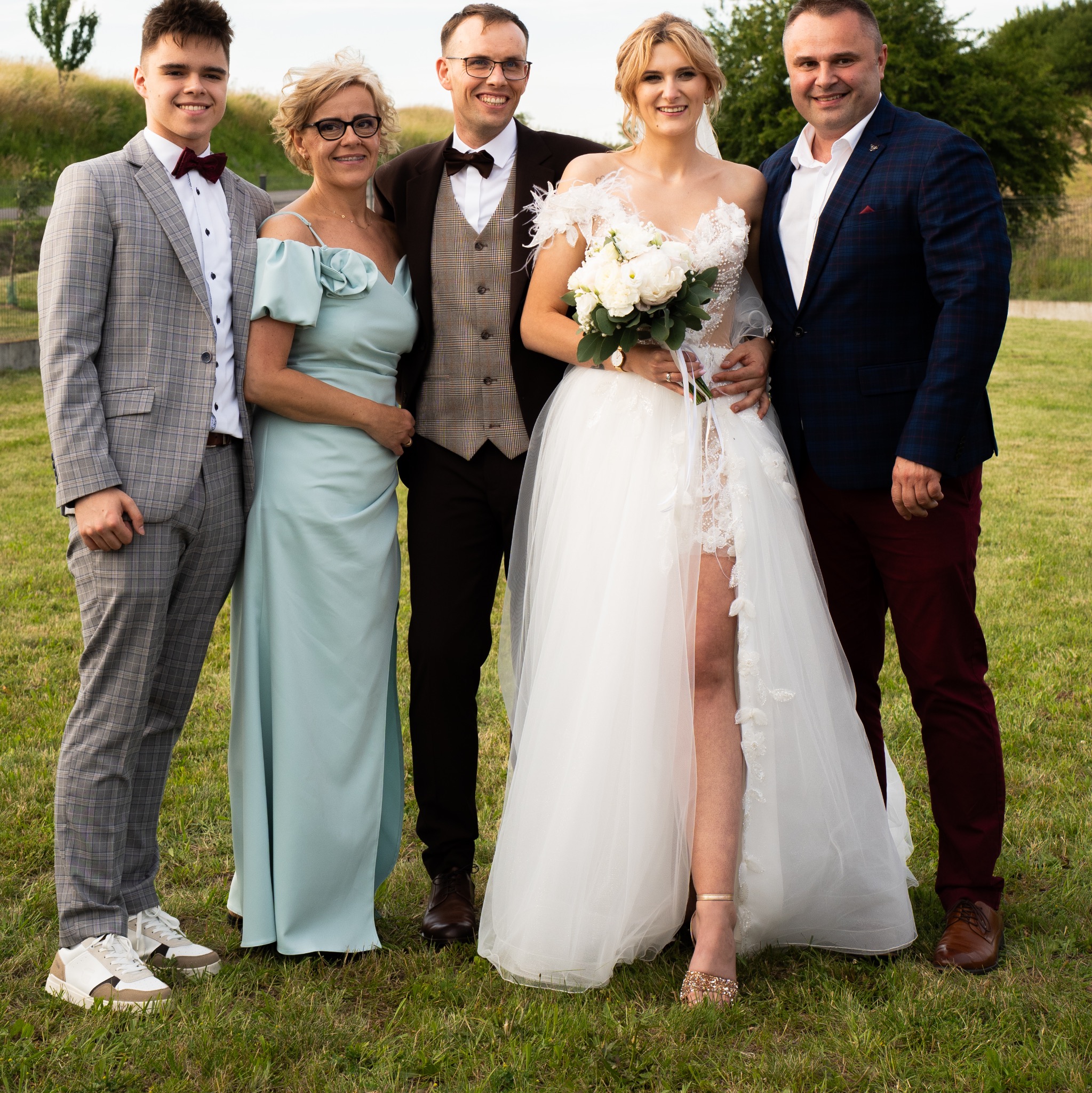 Uśmiechnięta panna młoda w koronkowej sukni z bukietem, otoczona elegancko ubraną rodziną na tle zielonej łąki w słoneczny dzień.