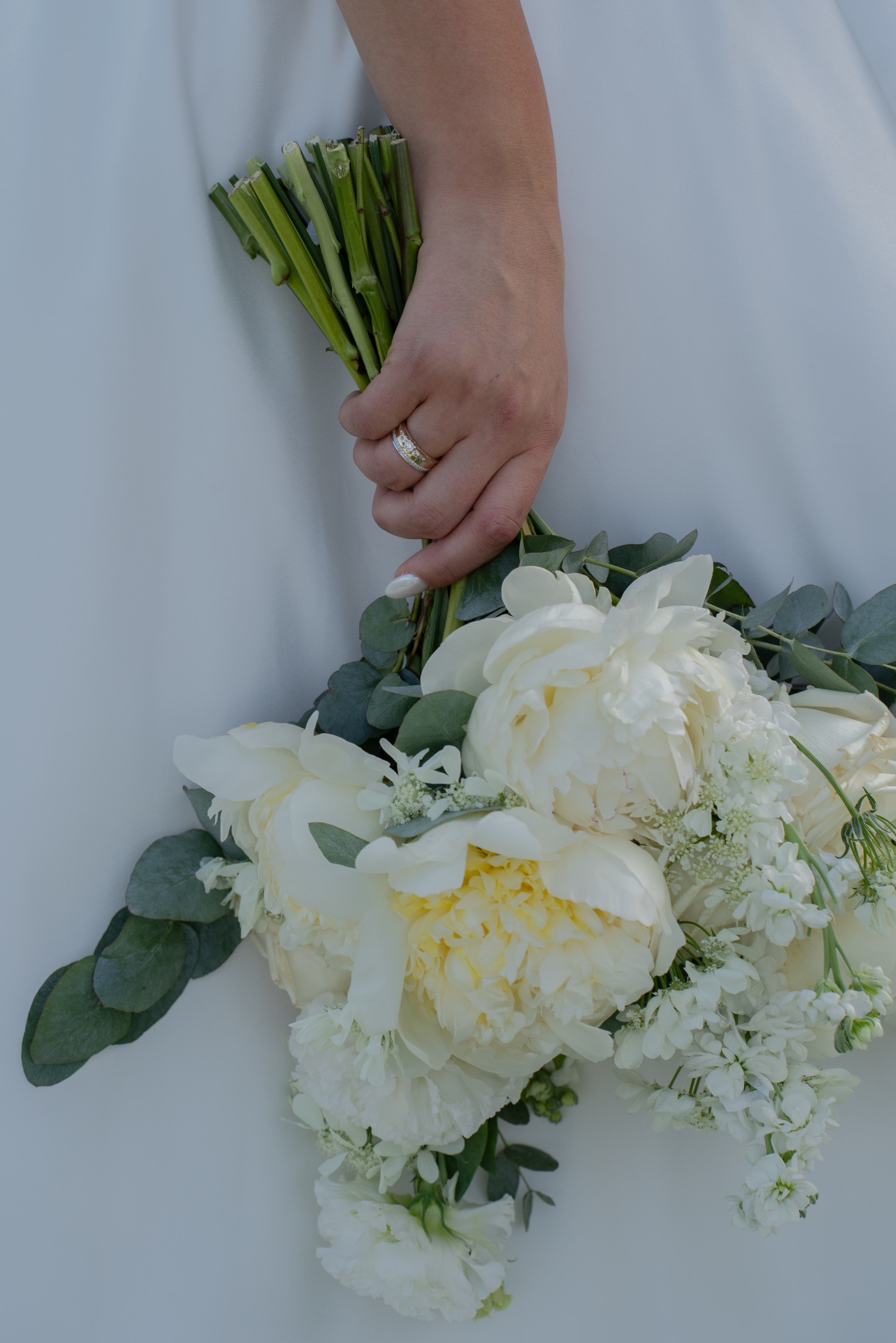 Dłoń z pierścionkiem trzyma bukiet ślubny z białych piwonii i eukaliptusa na tle białej sukni. Delikatne światło podkreśla detale kwiatów.