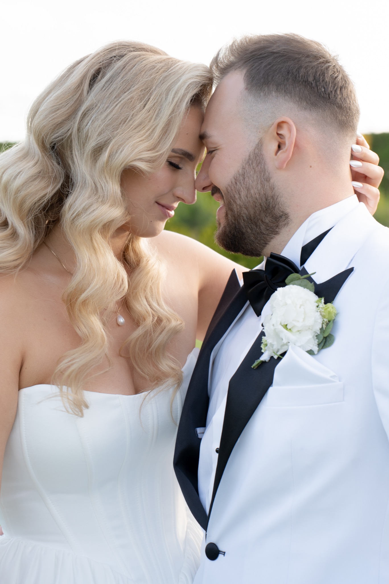 Uśmiechnięta para młoda, zbliżona twarzami w plenerze. Panna młoda w białej sukni, pan młody w białym smokingu z czarną muchą i butonierką.