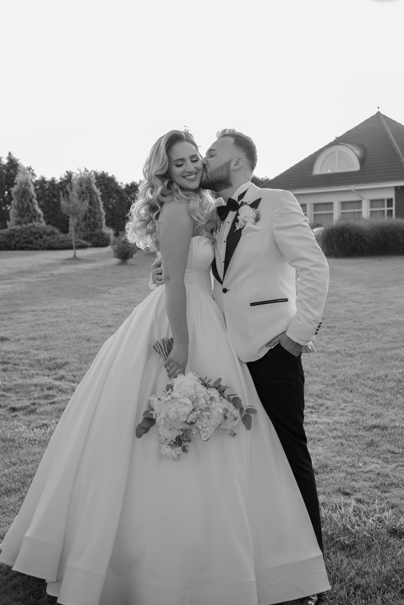 Czarno-białe zdjęcie całującej się pary młodej w plenerze na tle trawnika i domu. Panna młoda w sukni z bukietem, pan młody w smokingu.