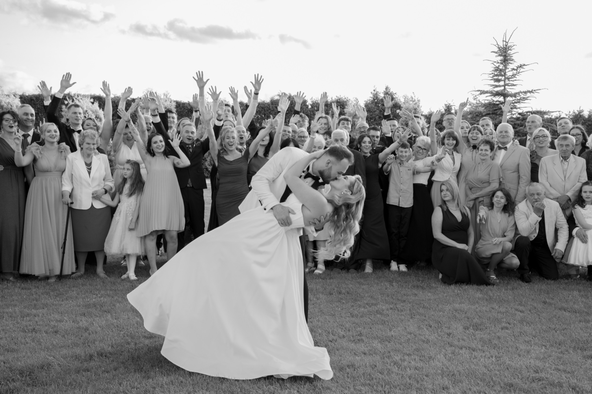 Czarno-białe zdjęcie całującej się pary młodej na tle wiwatujących gości weselnych na trawniku, z drzewami w tle. Dynamiczne ujęcie.