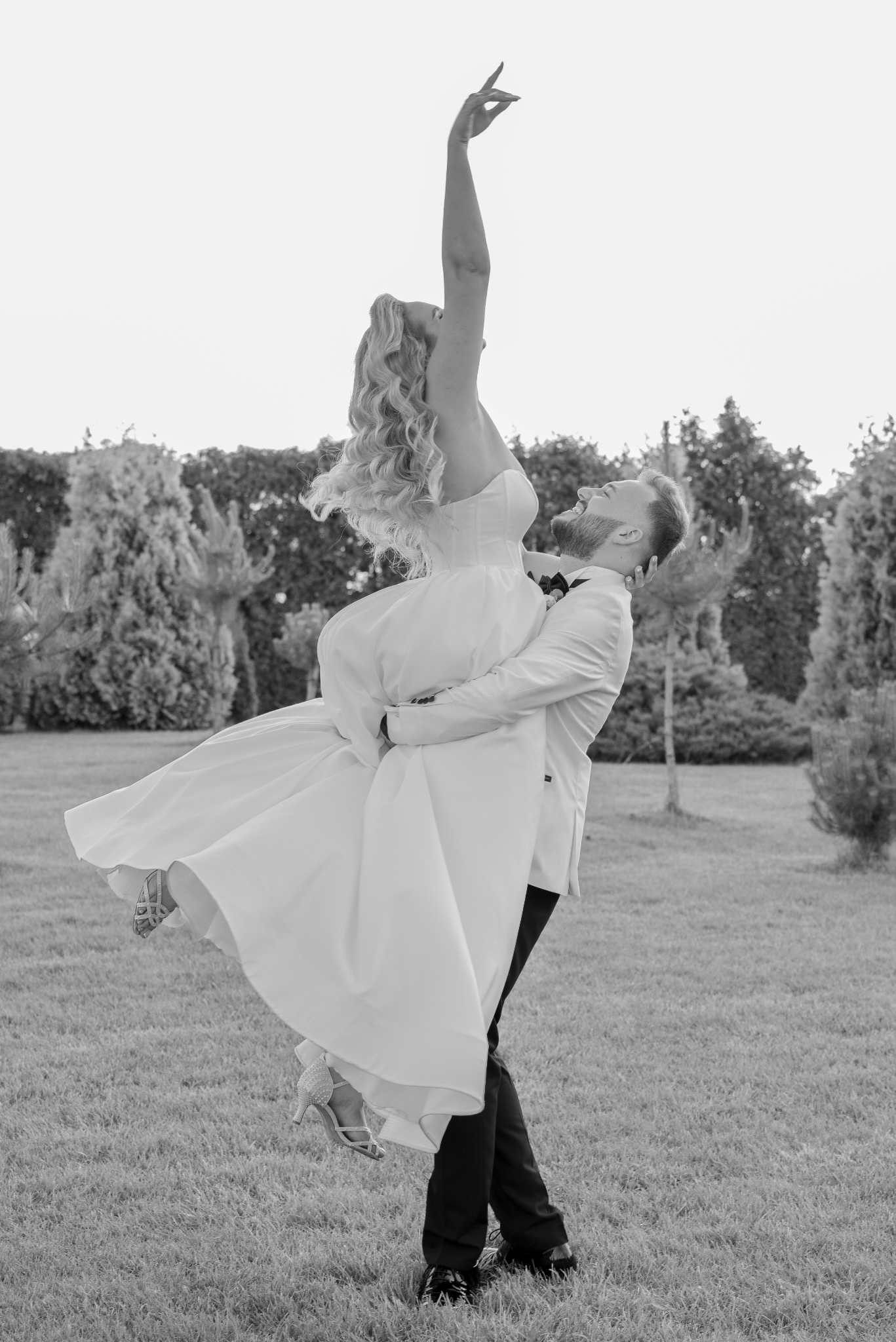 Czarno-białe zdjęcie: Pan młody unosi w górę uśmiechniętą pannę młodą w białej sukni, na tle zielonego ogrodu. Panna młoda z uniesioną ręką. Radosna atmosfera.