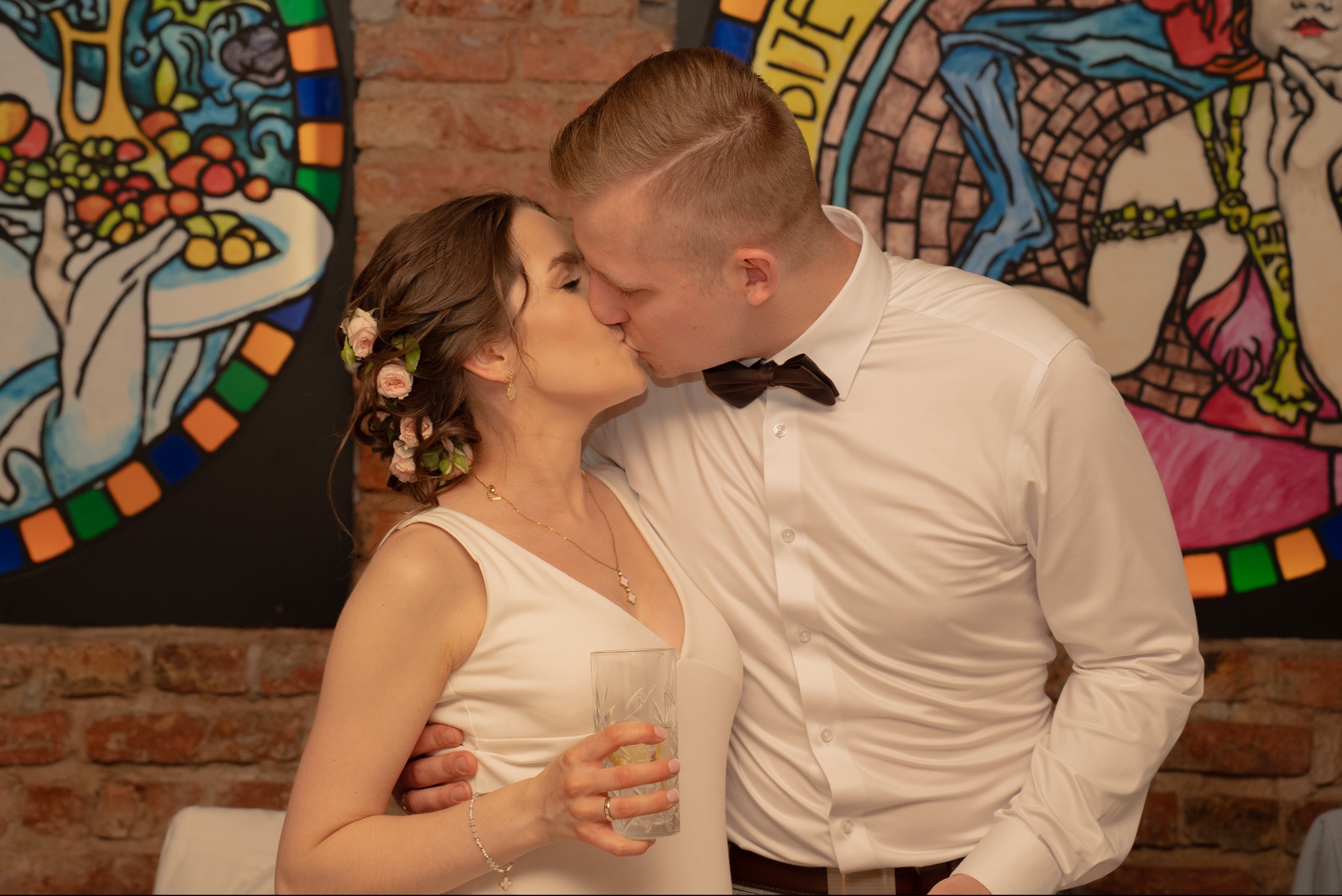Pocałunek młodej pary w jasnych strojach na tle ceglanej ściany z kolorową dekoracją. Panna młoda trzyma w dłoni kryształową szklankę.