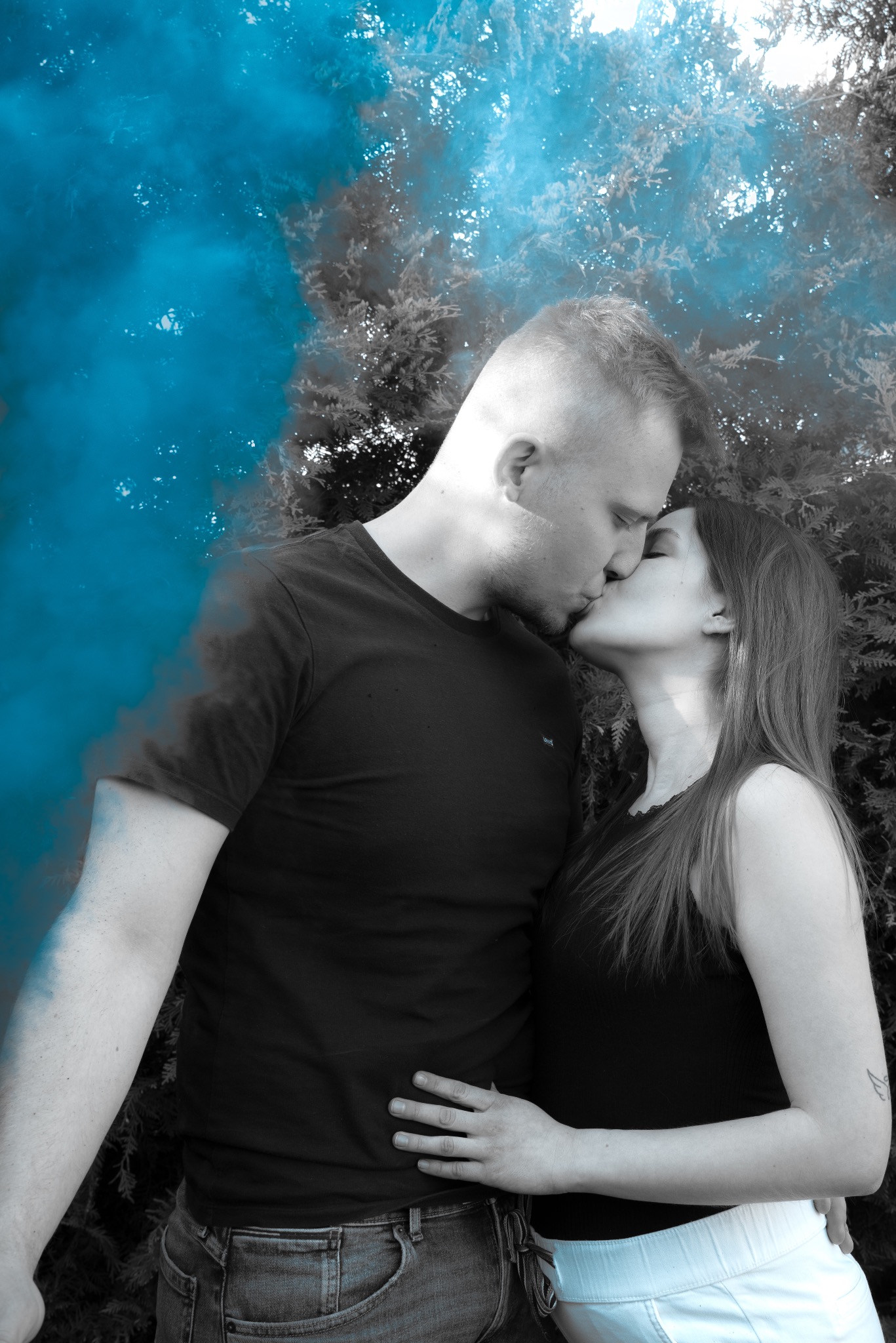 Para całująca się na tle drzew i niebieskiego dymu. Ujęcie z bliska, artystyczny efekt kolorystyczny, selektywna koloryzacja. Sesja plenerowa.