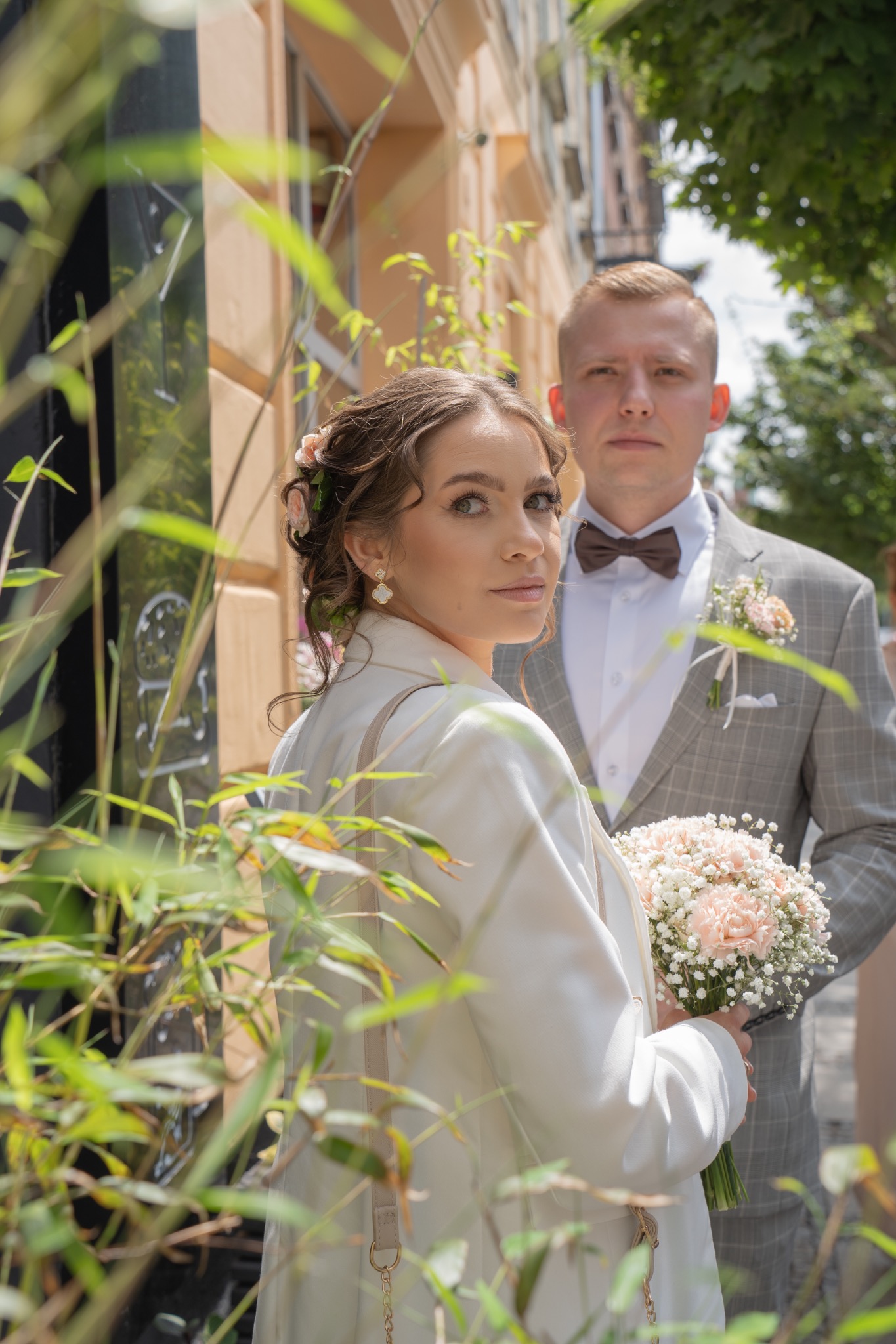 Para młoda w plenerze: panna młoda w białym żakiecie trzyma bukiet, obok pan młody w szarej marynarce z muszką. Ujęcie przez roślinność, jasne światło.