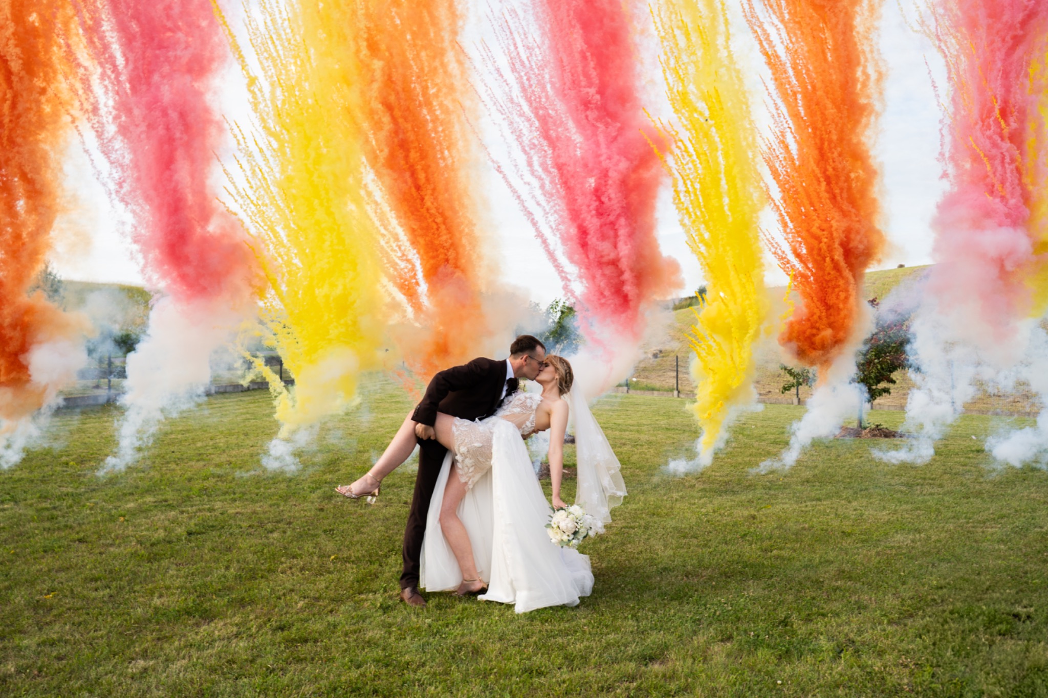 Para młoda w plenerze, mężczyzna trzyma kobietę w ramionach, w tle kolorowe pirotechniczne efekty specjalne w odcieniach żółci, pomarańczy i różu.