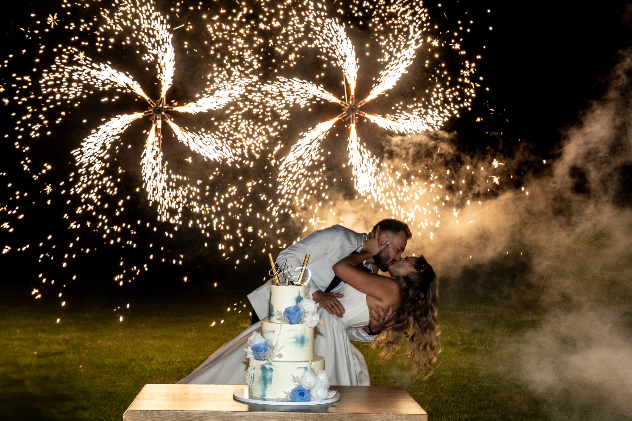 Pocałunek pary młodej na tle efektownych fajerwerków i tortu weselnego z dekoracjami kwiatowymi, uchwycony w kadrze fotografa ślubnego.