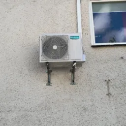 Zewnętrzna jednostka klimatyzacji Hisense zamontowana na wspornikach na szorstkiej, jasnej ścianie budynku, z widocznym fragmentem okna.