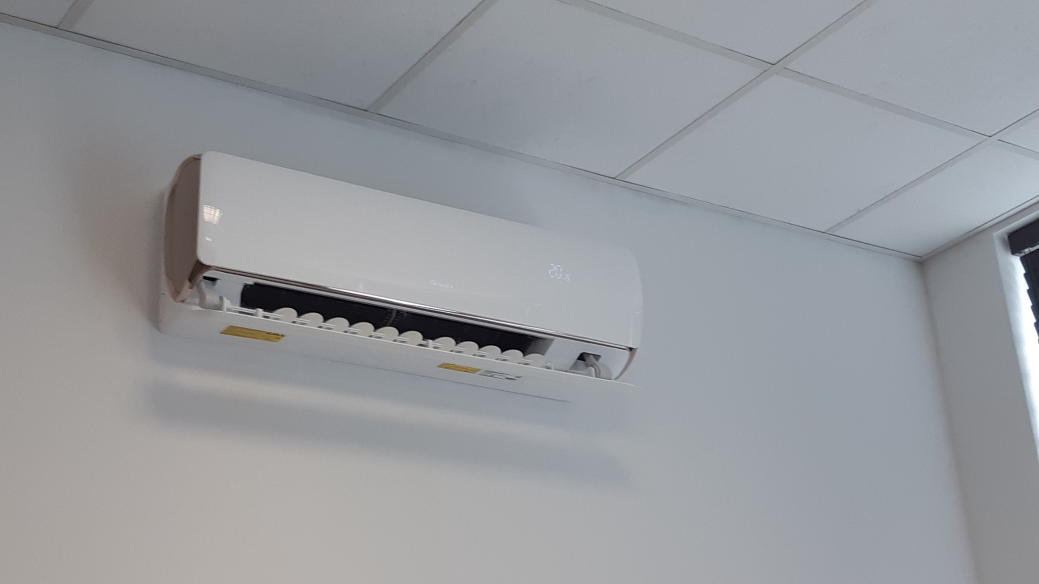 Biały klimatyzator ścienny marki Gree z wyświetlaczem temperatury 20.5 stopni Celsjusza, zamontowany w pomieszczeniu z kasetonowym sufitem.