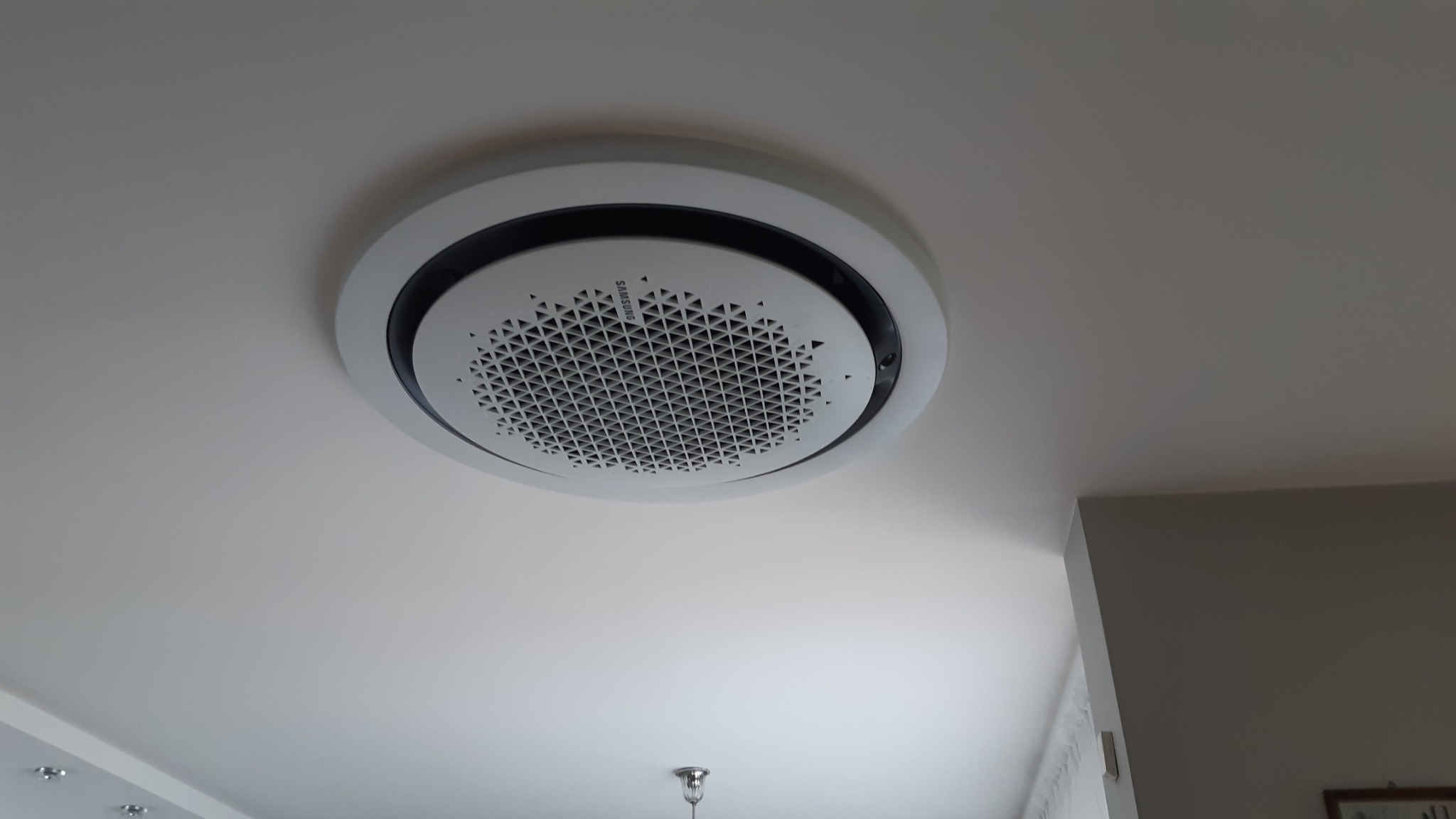 Sufitowy klimatyzator Samsung w okrągłej obudowie, zamontowany na białym suficie, widok z dołu.