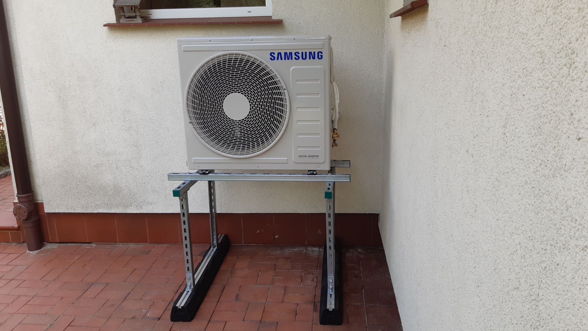 Zewnętrzna jednostka klimatyzacji Samsung Digital Inverter zamontowana na metalowej konstrukcji na tle ściany budynku z ceglaną podmurówką i brązową rynną.