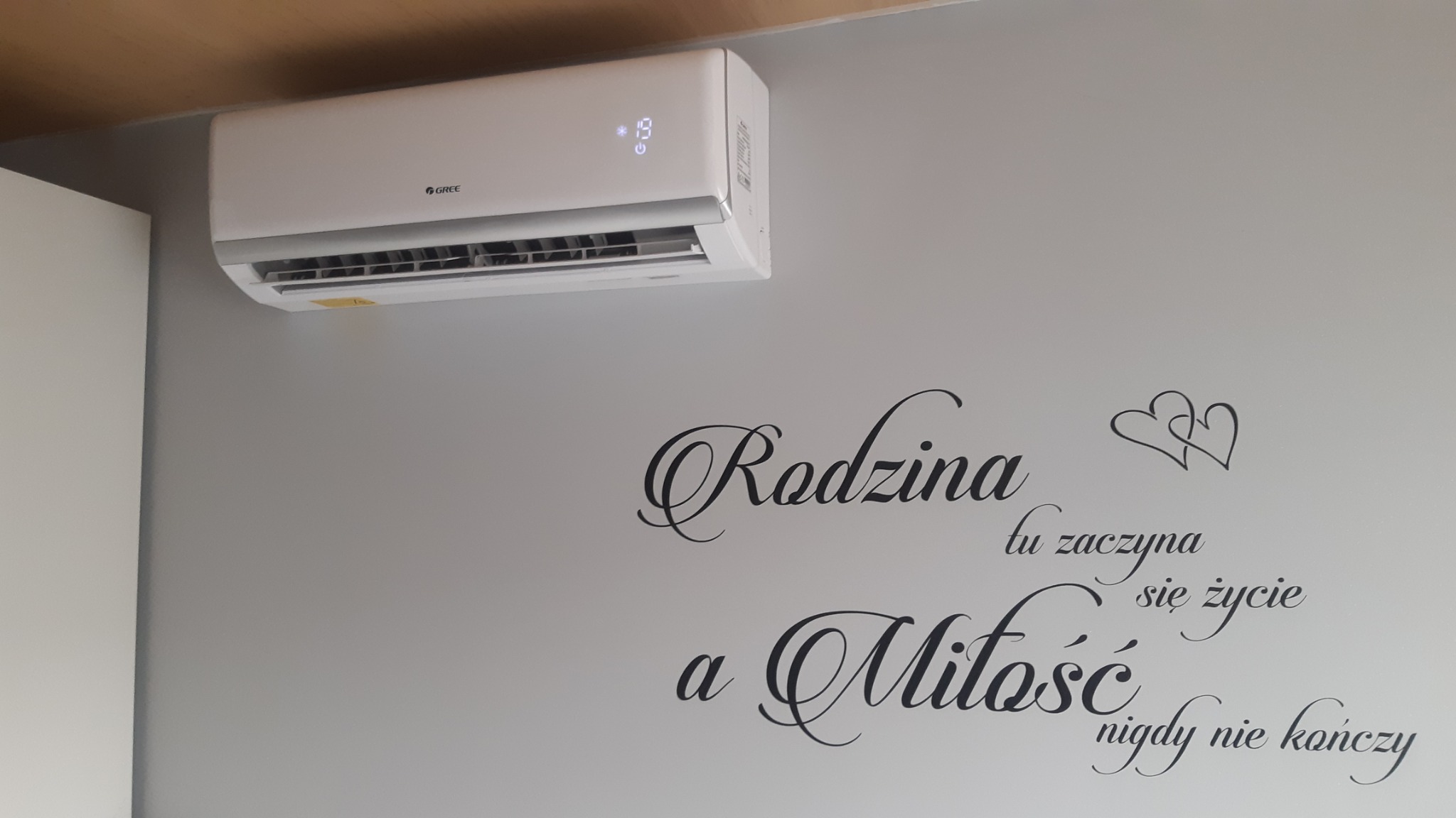 Biała klimatyzacja marki Gree zamontowana na szarej ścianie z dekoracyjnym napisem 'Rodzina tu zaczyna się życie a Miłość nigdy nie kończy' i dwoma sercami.
