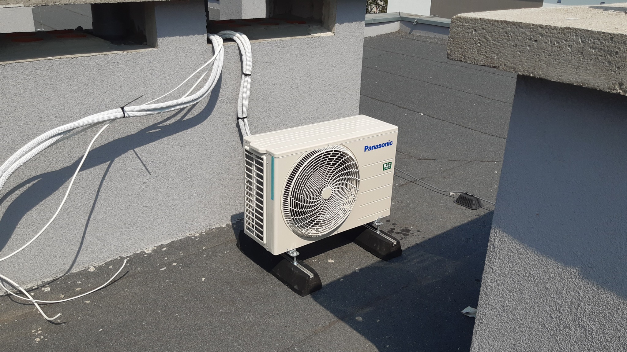 Zewnętrzna jednostka klimatyzacji Panasonic zamontowana na płaskim dachu budynku, z widocznymi przewodami zabezpieczonymi peszlami i przymocowanymi do ściany.