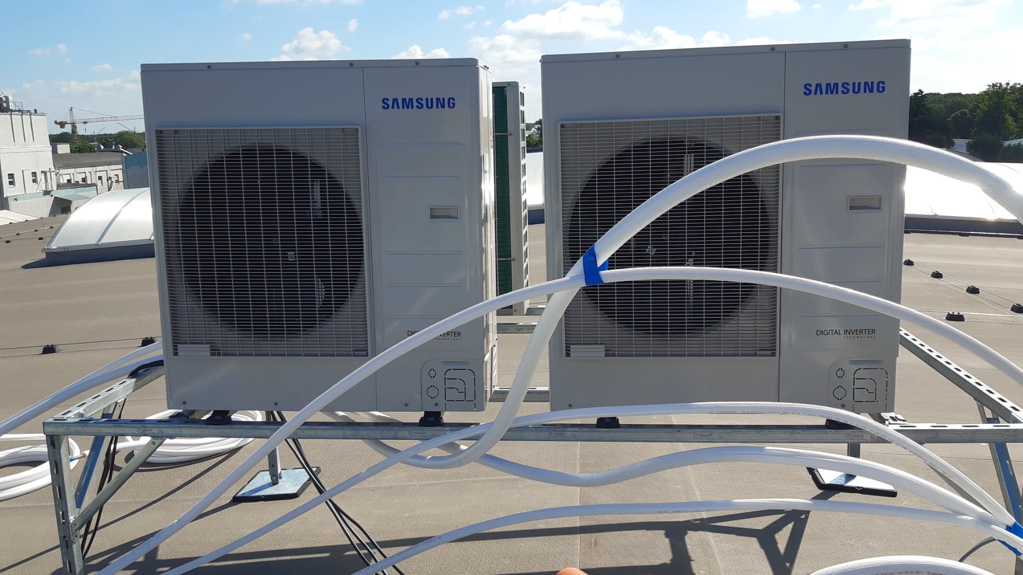 Dwa zewnętrzne moduły klimatyzacji Samsung Digital Inverter na dachu budynku, połączone białymi rurami, zamocowane na metalowej konstrukcji.