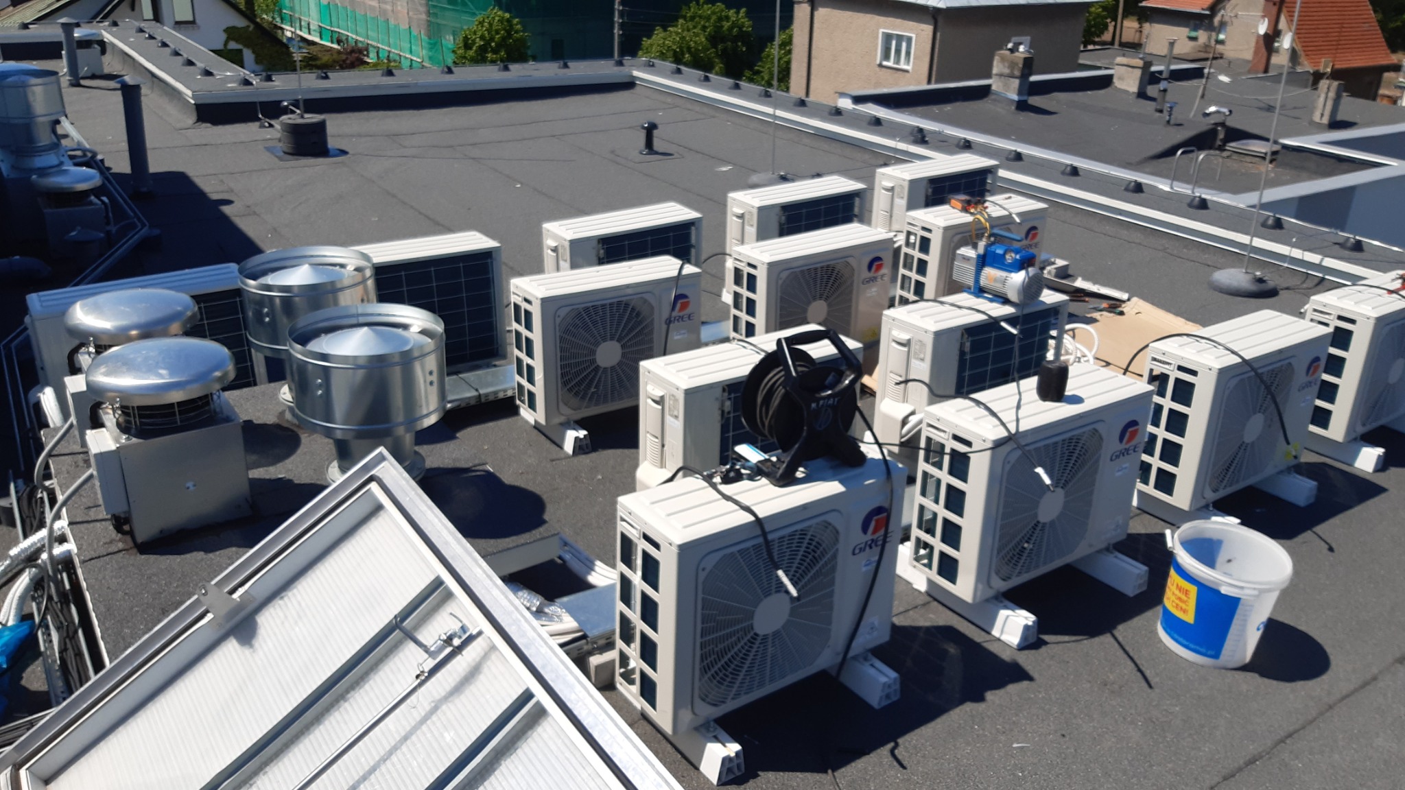 Instalacja klimatyzacji na dachu budynku, widoczne jednostki zewnętrzne klimatyzatorów marki Gree, wentylatory dachowe, narzędzia pracy oraz wiadro z niebieskim paskiem.