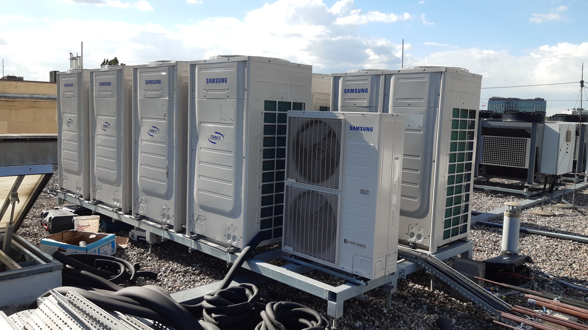 Rząd zewnętrznych jednostek klimatyzacyjnych Samsung DVM S na dachu budynku, widoczne elementy instalacji i rurki miedziane.