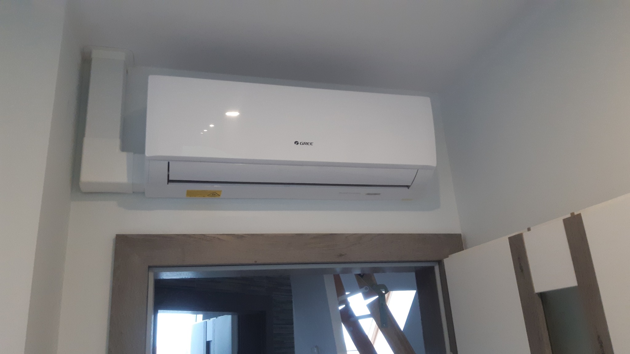 Biały klimatyzator marki Gree zamontowany na ścianie w pomieszczeniu z widocznym fragmentem futryny drzwi i częścią zabudowy meblowej.
