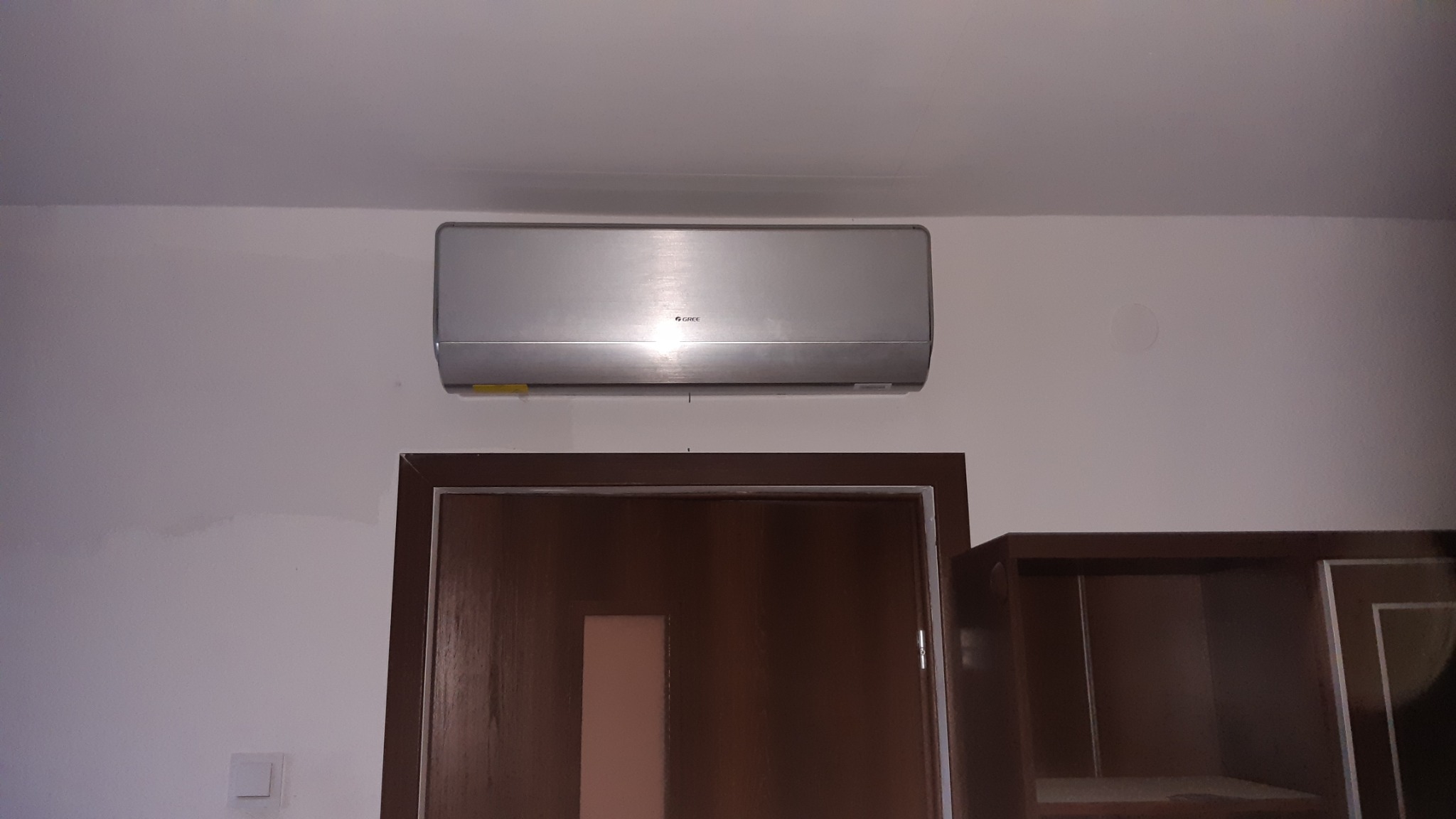 Srebrny klimatyzator marki Gree zamontowany na białej ścianie nad brązowymi drzwiami i częścią szafki.