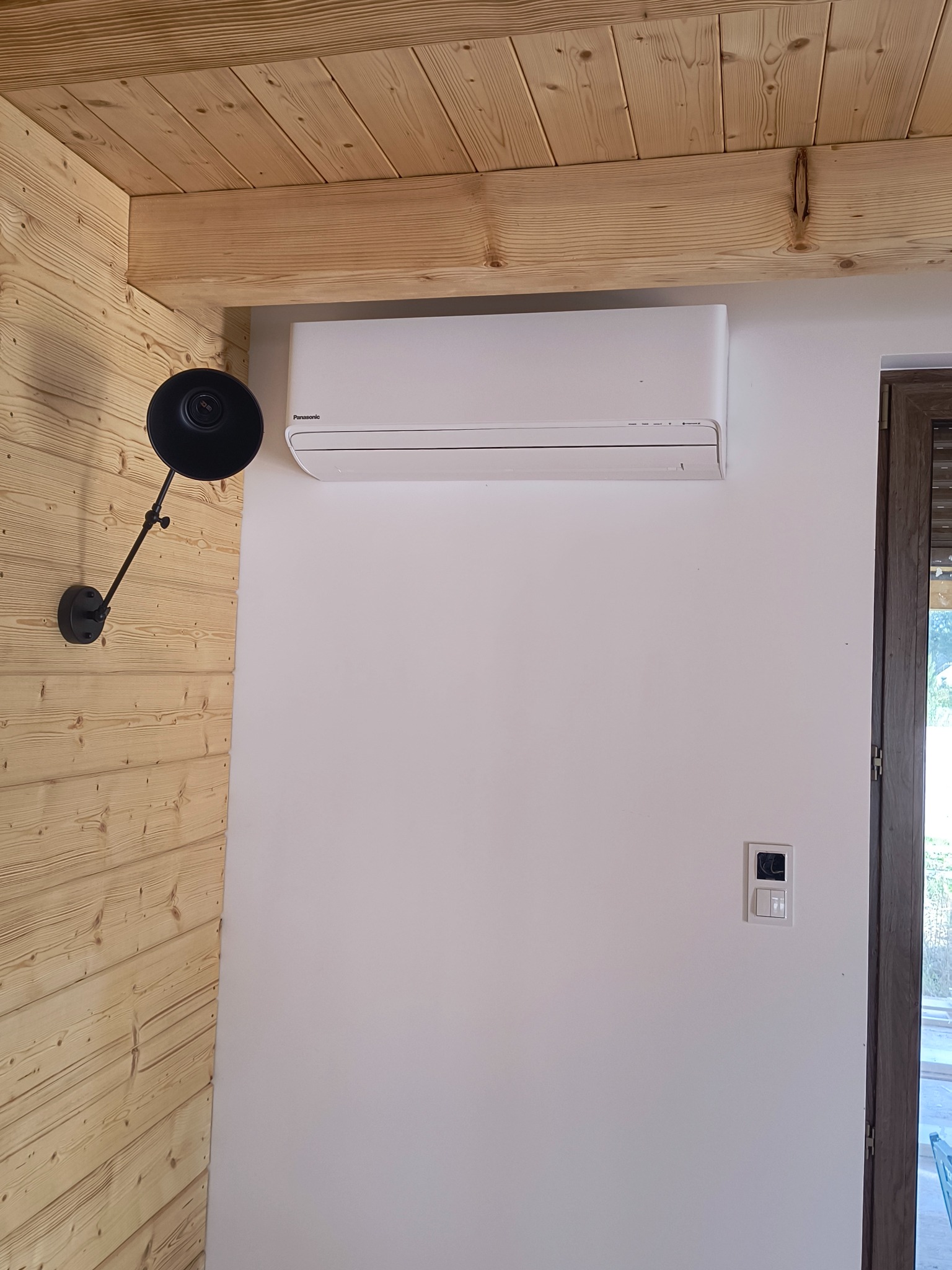 Biały klimatyzator Panasonic zamontowany na białej ścianie w pomieszczeniu z drewnianą boazerią i lampą ścienną