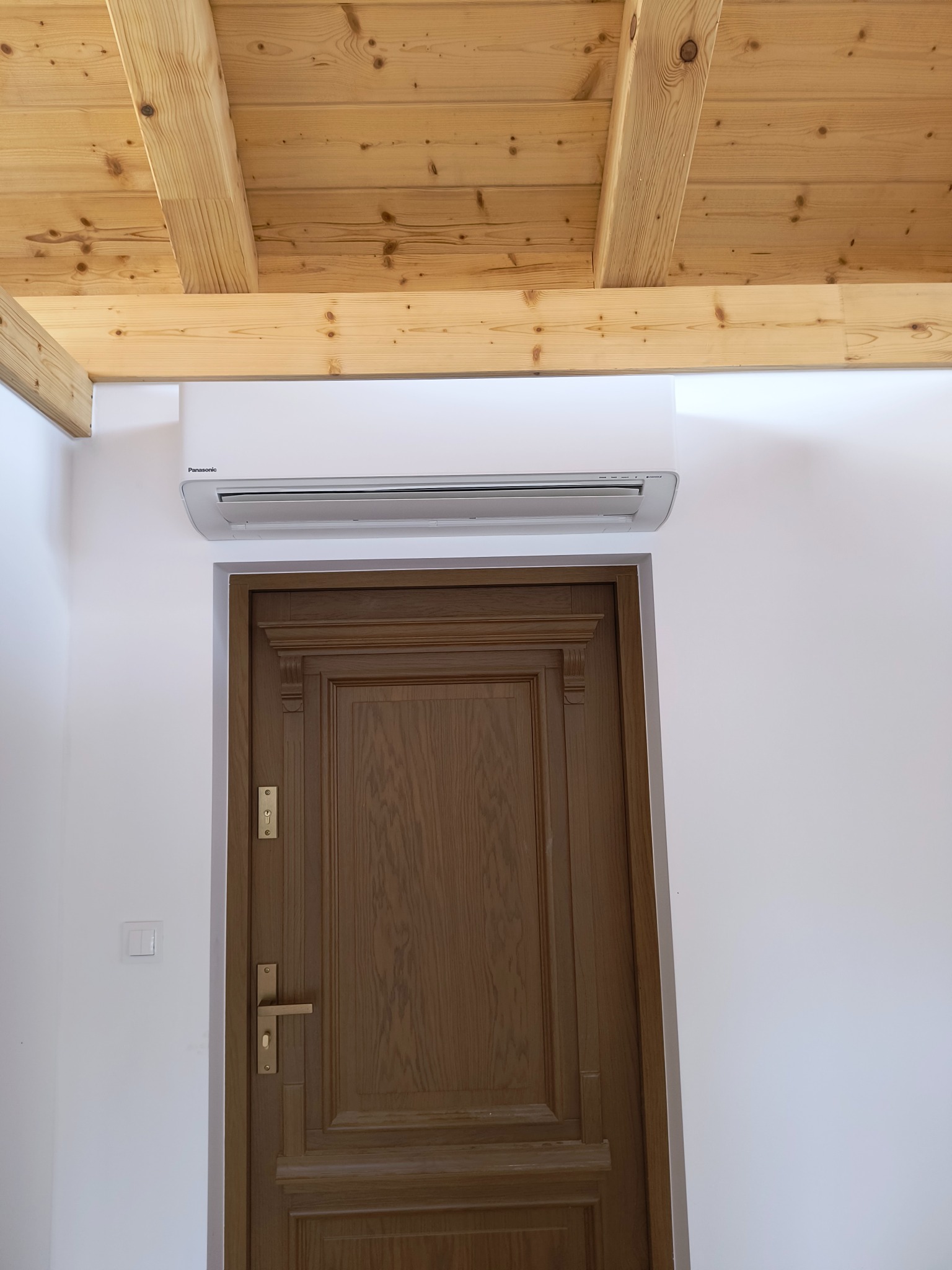 Biała jednostka klimatyzacyjna Panasonic zamontowana nad drewnianymi drzwiami w białym pomieszczeniu z drewnianym sufitem