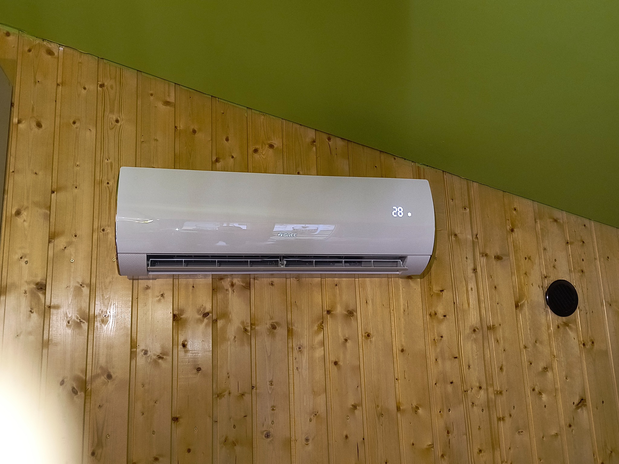 Biały klimatyzator marki Gree zamontowany na ścianie wyłożonej boazerią, z wyświetlaczem wskazującym temperaturę 28 stopni Celsjusza.