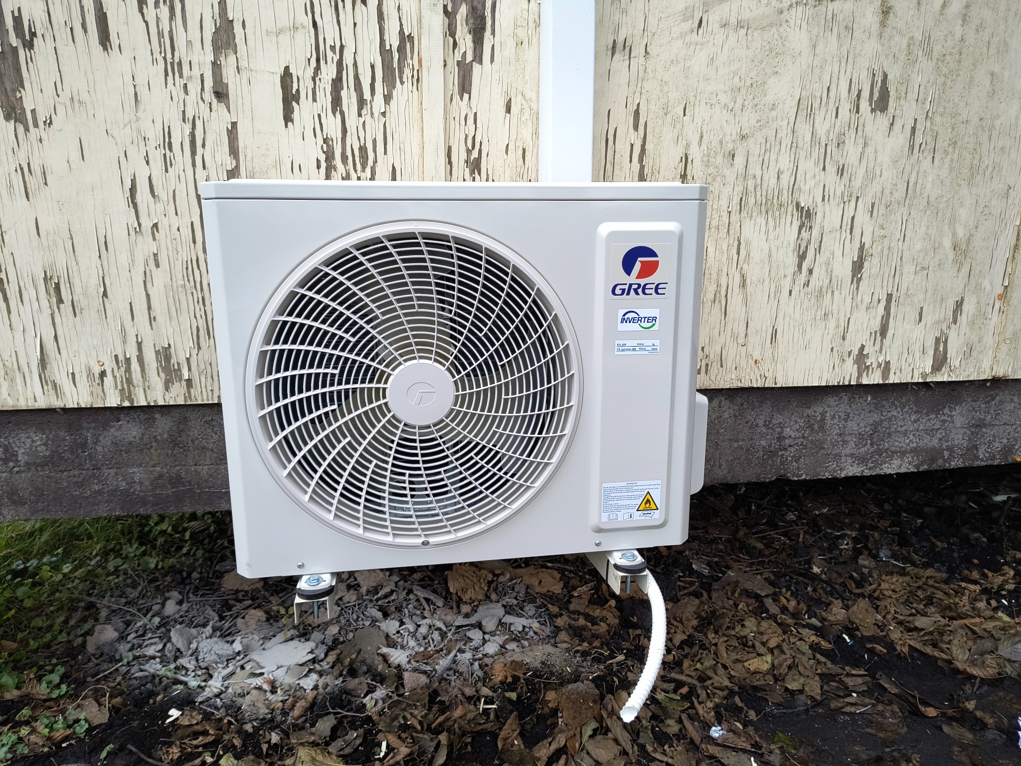 Zewnętrzna jednostka klimatyzacji marki Gree zamontowana na ścianie budynku z łuszczącą się farbą, widoczne mocowania i rura odprowadzająca.