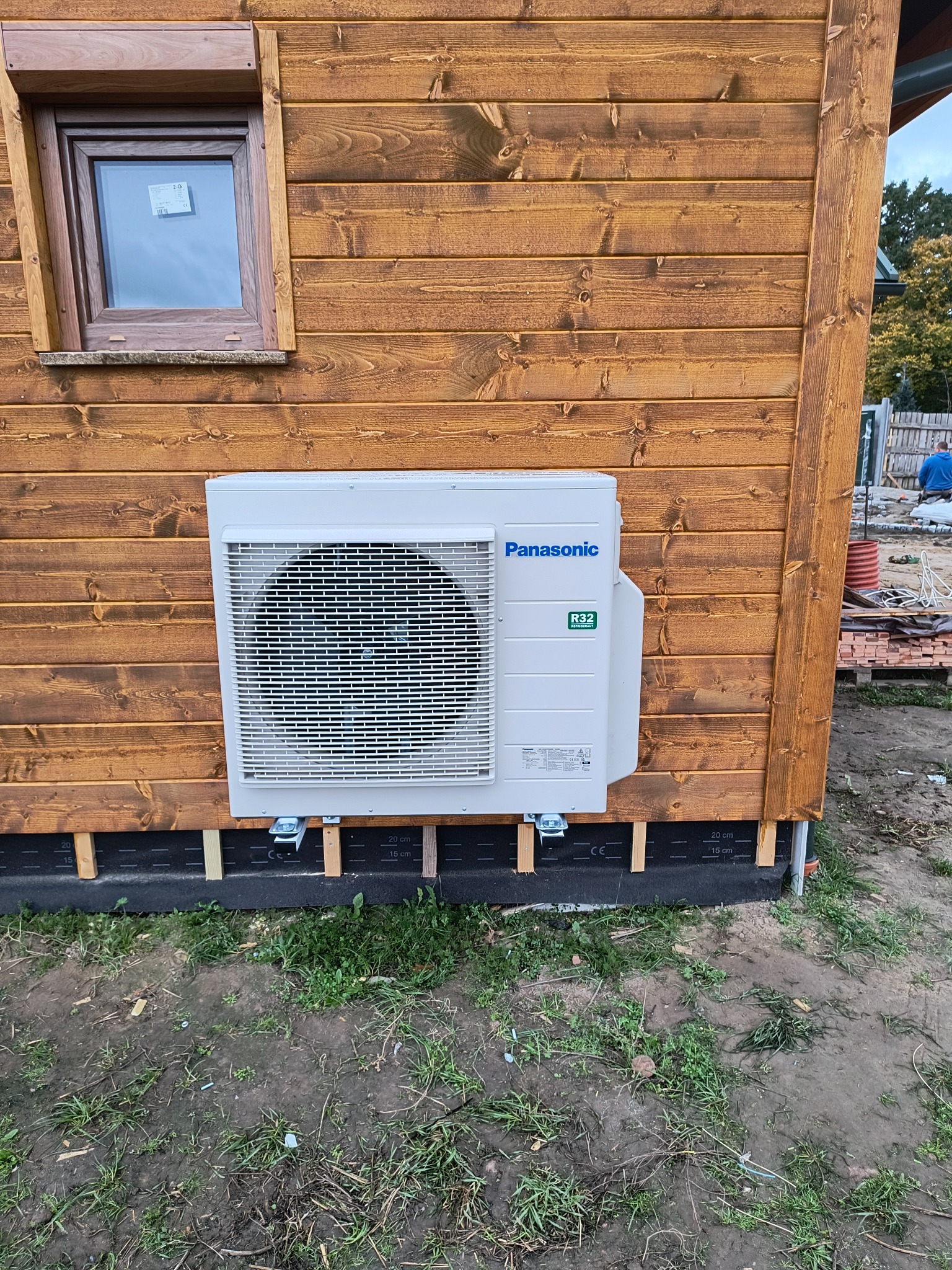 Biała jednostka zewnętrzna klimatyzacji Panasonic R32 zamontowana na drewnianej ścianie budynku z widocznym oknem i elementami konstrukcyjnymi na dole, teren wokół częściowo trawiasty, częściowo...