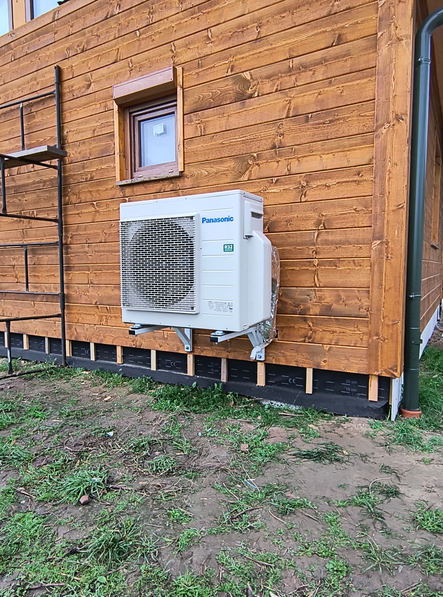 Zewnętrzna jednostka klimatyzacji Panasonic zamontowana na drewnianej elewacji domu, widoczna drabina i małe okno.