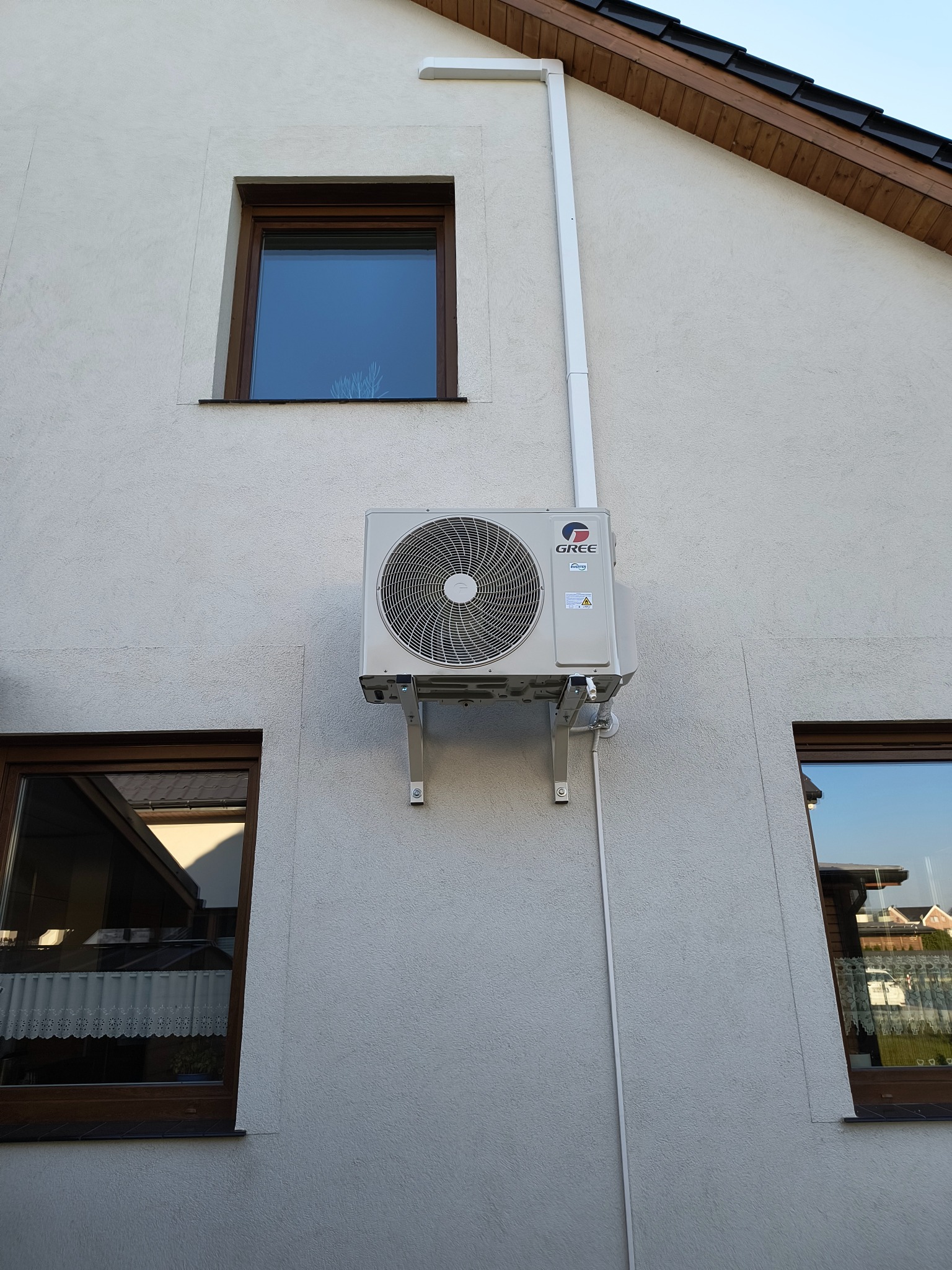 Zewnętrzna jednostka klimatyzacji marki Gree zamontowana na elewacji domu, widoczne okna z drewnianą ramą i fragment dachu.