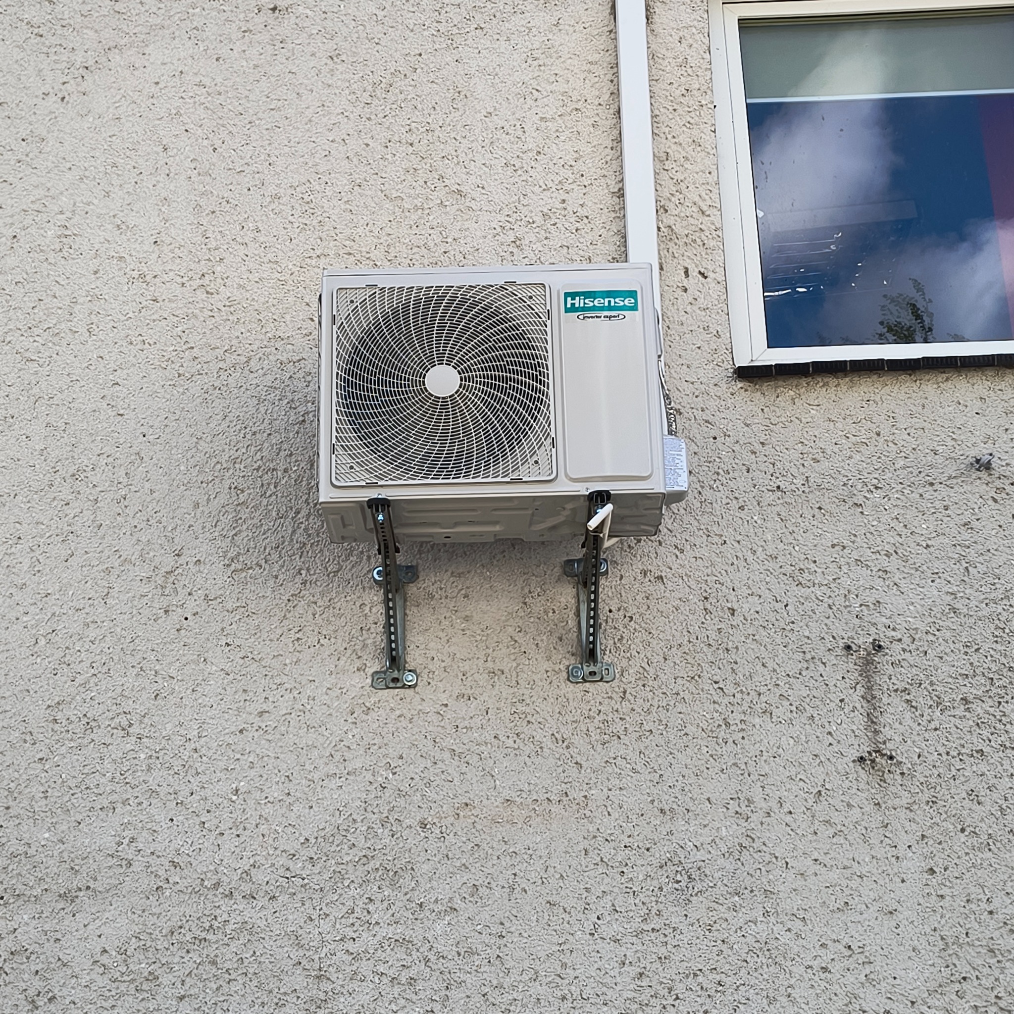 Zewnętrzna jednostka klimatyzacji Hisense zamontowana na wspornikach na szorstkiej, jasnej ścianie budynku, z widocznym fragmentem okna.