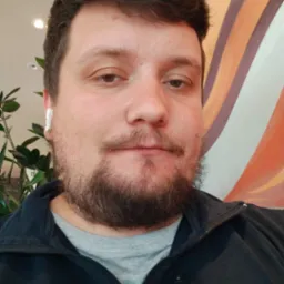 Mężczyzna z brodą i słuchawką bezprzewodową w uchu, ubrany w czarną bluzę, na tle ściany w kolorowe pasy i rośliny doniczkowej.