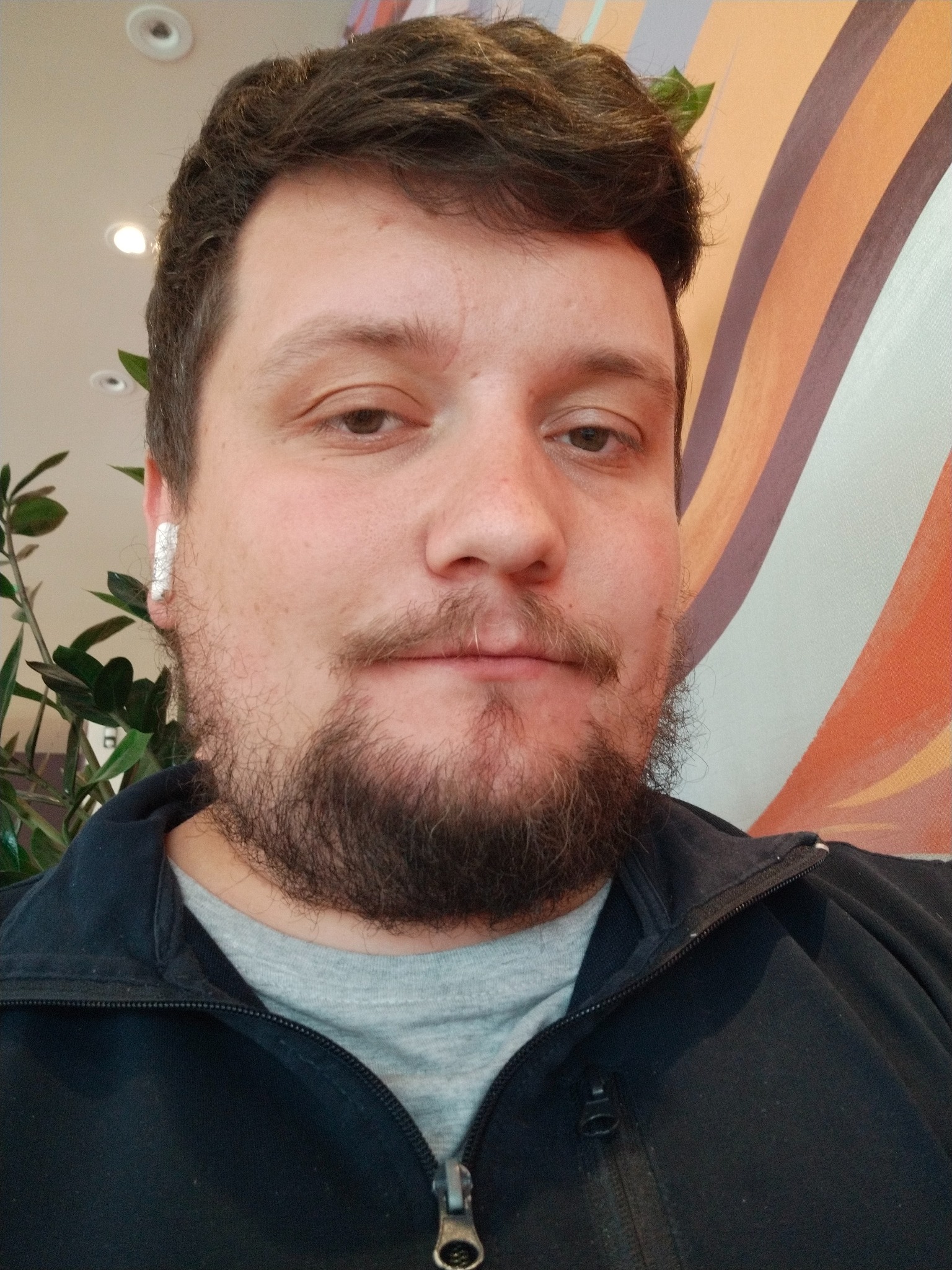 Mężczyzna z brodą i słuchawką bezprzewodową w uchu, ubrany w czarną bluzę, na tle ściany w kolorowe pasy i rośliny doniczkowej.