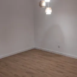 Puste pomieszczenie z nowo ułożonymi panelami podłogowymi w odcieniach brązu, oświetlone przez designerską lampę wiszącą z abażurami z naturalnego materiału.