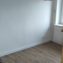 Vian - Pusty pokój po remoncie z drewnopodobną podłogą, świeżo pomalowanymi białymi ścianami, dekoracyjną lampą wiszącą i grzejnikiem pod oknem.