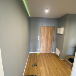 Vian - Szare ściany, drewnopodobna podłoga i drzwi wejściowe w wyremontowanym przedpokoju. Widoczne narzędzia i oświetlenie LED na suficie. Ujęcie z dołu.