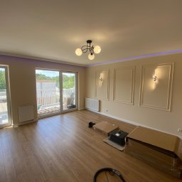 Vian - Jasne wnętrze po remoncie: drewniana podłoga, ozdobne listwy na ścianach z oświetleniem, balkon z widokiem na zieleń. Częściowo rozpakowane elementy wyposażenia.