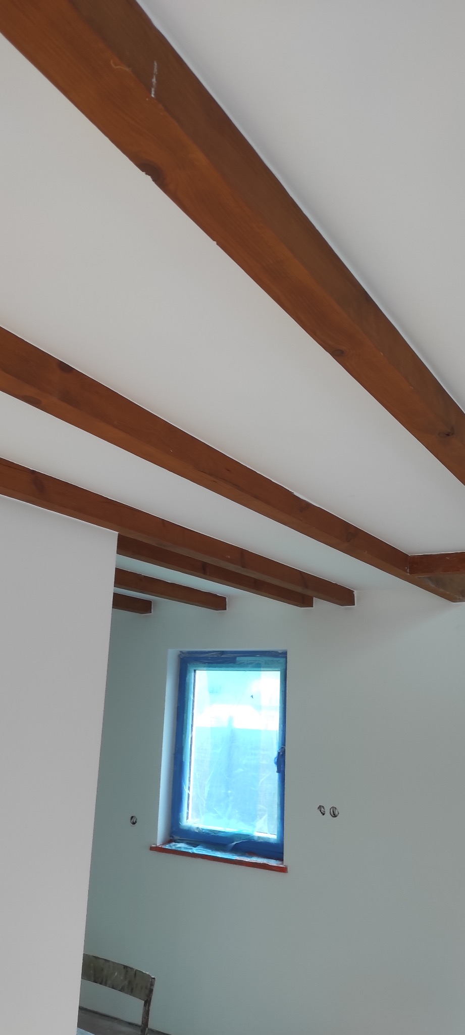 Wnętrze pokoju w trakcie malowania: białe ściany, drewniane belki stropowe, okno zabezpieczone folią malarską.