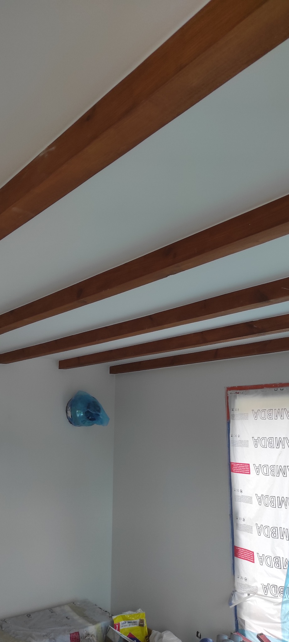 Białe wnętrze z drewnianymi belkami stropowymi, okno zabezpieczone folią ochronną, grzejnik osłonięty niebieską folią, widoczne materiały budowlane na podłodze.