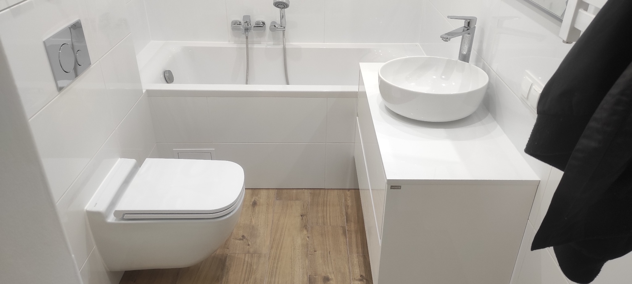 Biała, nowoczesna łazienka z wiszącą miską WC, okrągłą umywalką nablatową i wanną w zabudowie, wykończona białymi płytkami i panelami podłogowymi imitującymi drewno.