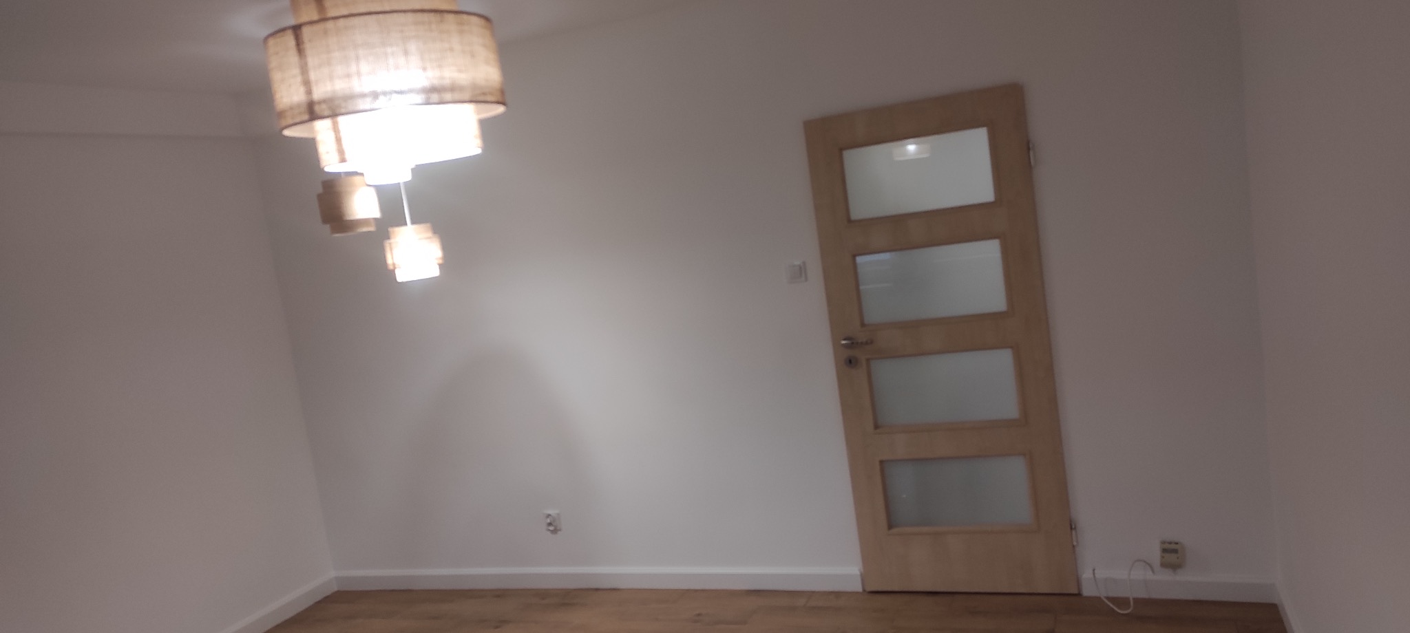 Jasne wnętrze z drewnianą podłogą, białymi ścianami i drzwiami z przeszkleniami; widoczna lampa sufitowa z abażurem.