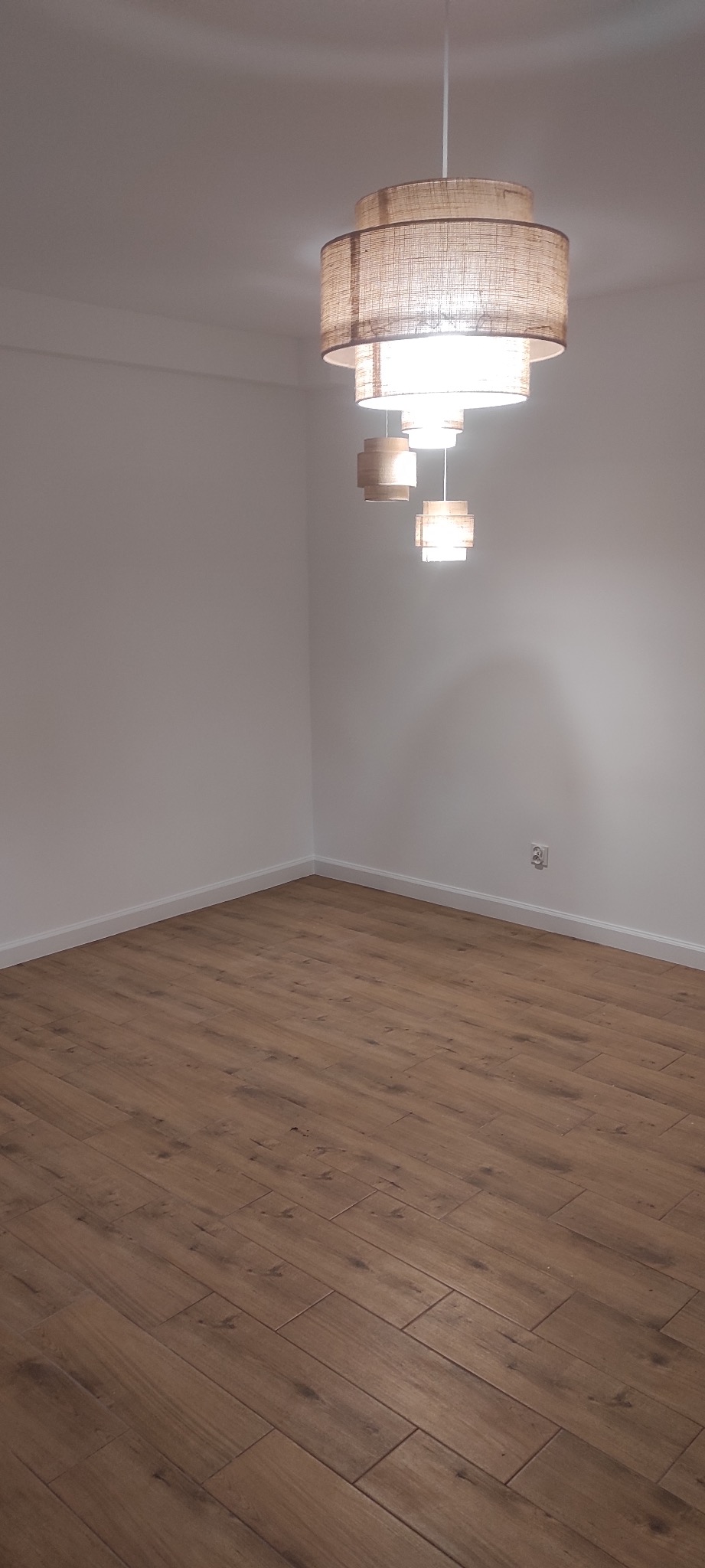 Puste pomieszczenie z nowo ułożonymi panelami podłogowymi w odcieniach brązu, oświetlone przez designerską lampę wiszącą z abażurami z naturalnego materiału.