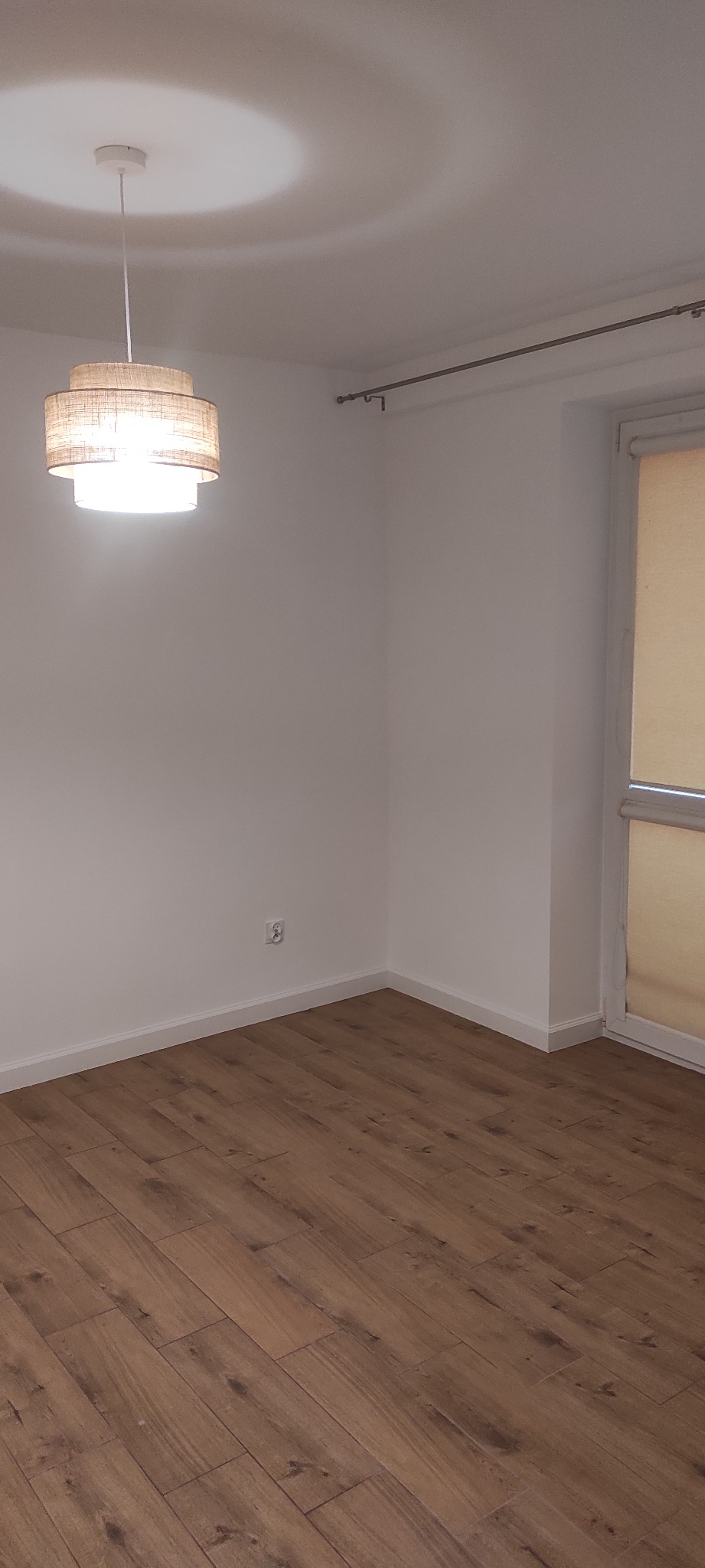 Pusty pokój z panelami podłogowymi w odcieniach brązu, białymi ścianami, oświetlony wiszącą lampą z abażurem z naturalnego materiału i światłem dziennym wpadającym przez okno z roletą.