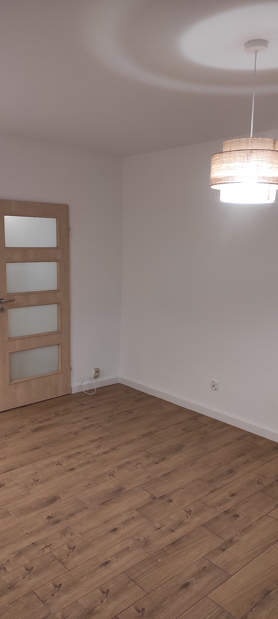 Puste, odnowione wnętrze w Świnoujściu z drewnopodobną podłogą, białymi ścianami i nowoczesną lampą sufitową. Widoczna jasna, przeszklona stolarka drzwiowa.