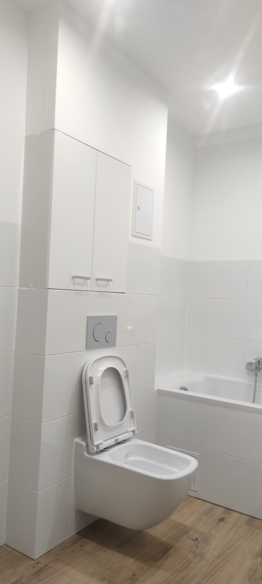 Nowoczesna, biała łazienka z wiszącą toaletą z otwartą deską, zabudową nad toaletą i wanną w zabudowie z białymi płytkami, wykończona podłogą imitującą drewno.