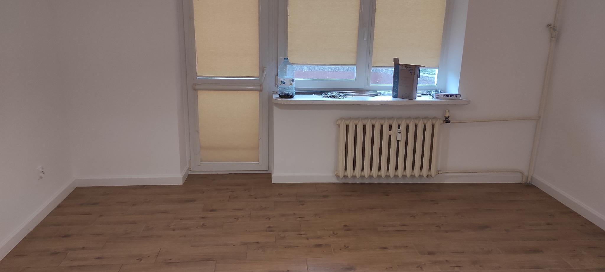 Pusty pokój po remoncie z nowymi panelami podłogowymi, świeżo pomalowanymi ścianami i starym grzejnikiem pod oknem, na parapecie butelka wody i pudełko. Widoczne ślady po gniazdkach elektrycznych.