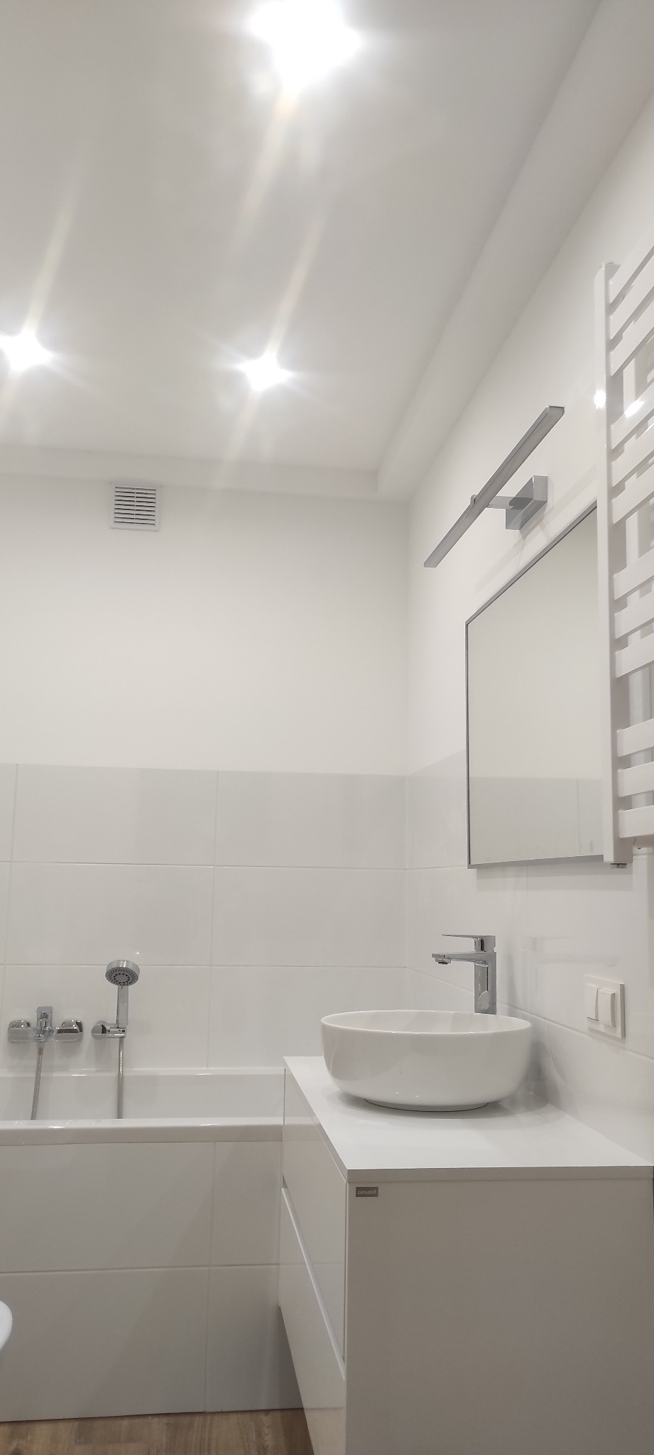 Jasna łazienka po remoncie: białe płytki, wanna z baterią, umywalka nablatowa na szafce z logo Cersanit, grzejnik drabinkowy i oświetlenie LED nad lustrem.