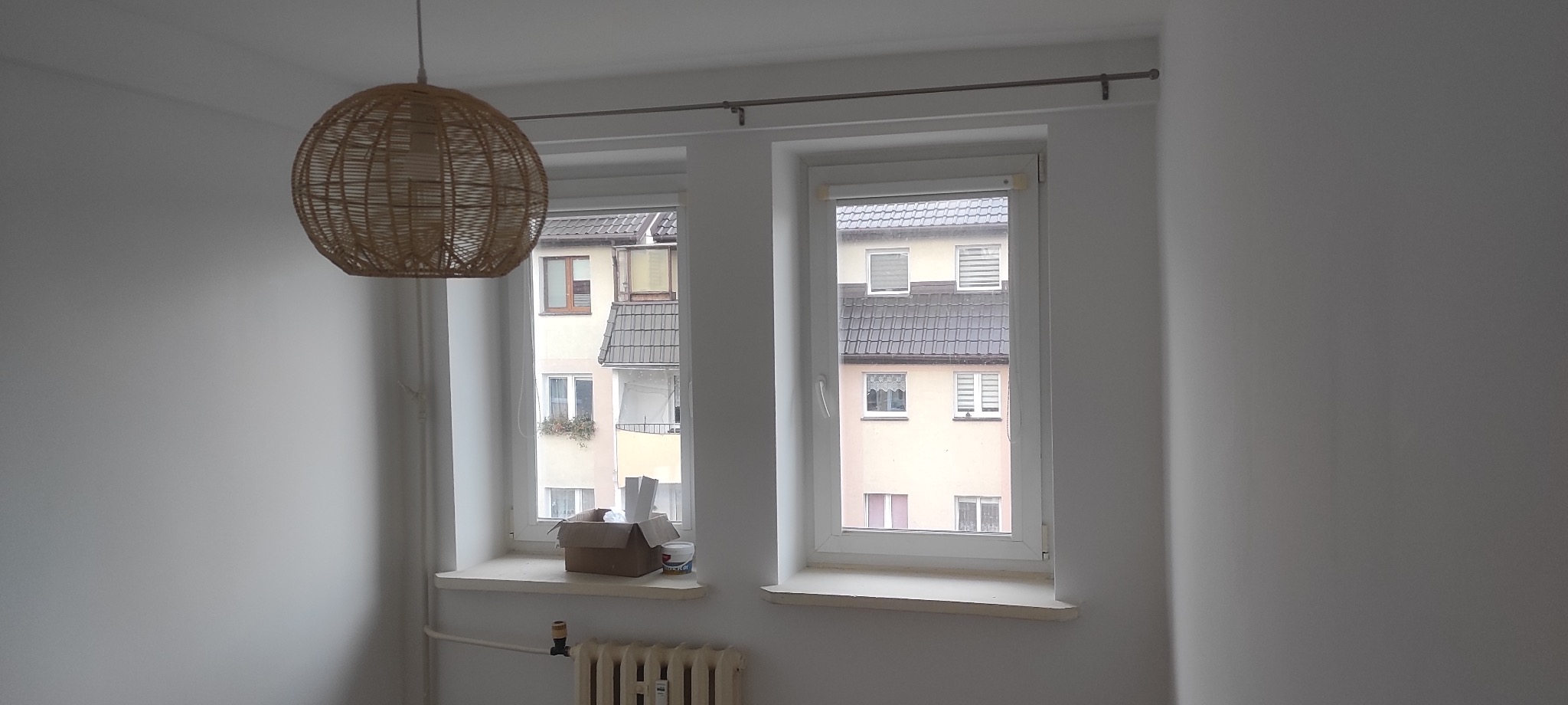 Wnętrze pokoju z dwoma oknami po remoncie, na parapetach karton i puszka z farbą, w tle widok na budynki, w górnej części kadru pleciona, okrągła lampa.