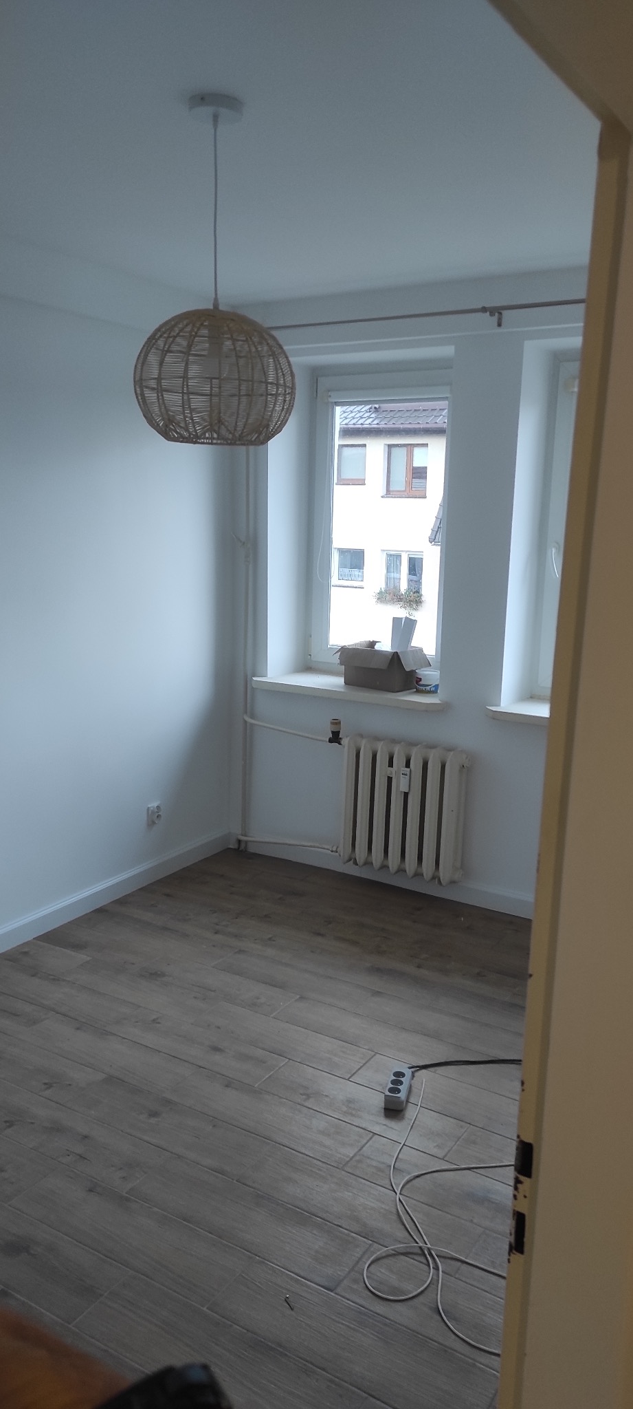 Pusty pokój po świeżym remoncie z drewnopodobną podłogą, białymi ścianami, grzejnikiem pod oknem i lampą z abażurem z wikliny, z widoczną listwą przypodłogową i gniazdkiem elektrycznym.
